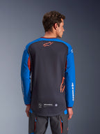 Maillot Alpinestars Maxdura Dual en gris foncé et bleu royal, vue de dos d'un modèle masculin, torse en maille avec logo Astars orange, manches longues avec marquage blanc, col et poignets bordeaux, porté avec un pantalon technique gris, équipement de moto dual-sport et d'aventure