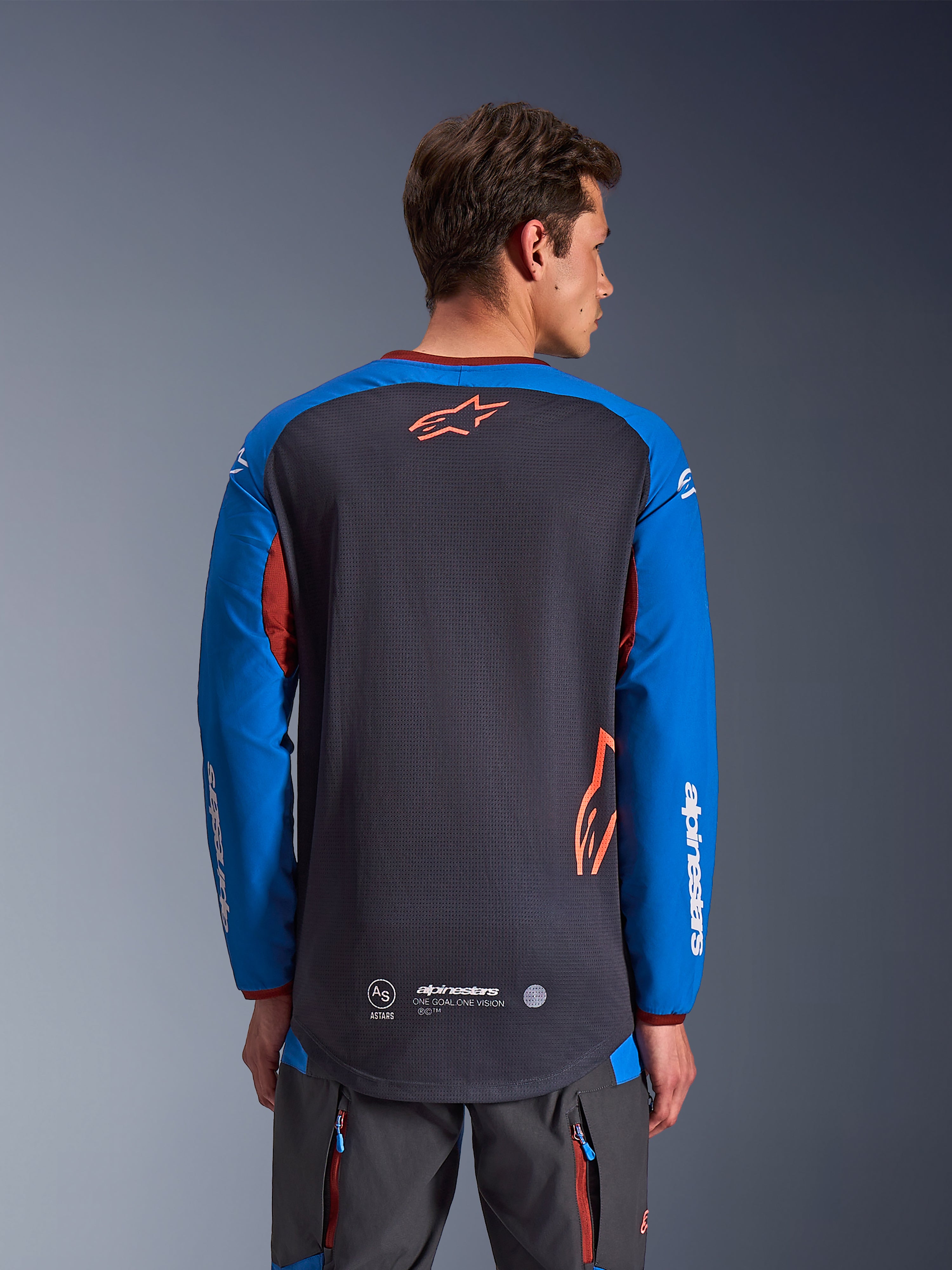 Maillot Alpinestars Maxdura Dual en gris foncé et bleu royal, vue de dos d'un modèle masculin, torse en maille avec logo Astars orange, manches longues avec marquage blanc, col et poignets bordeaux, porté avec un pantalon technique gris, équipement de moto dual-sport et d'aventure