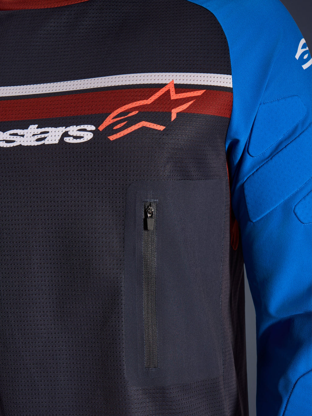 Maillot Alpinestars Maxdura Dual, gros plan sur la poitrine et la manche, gris foncé et bleu royal, avec poche poitrine zippée, torse en maille respirante et rembourrage intégré sur les manches, logo Alpinestars sur bandes rouges et blanches
