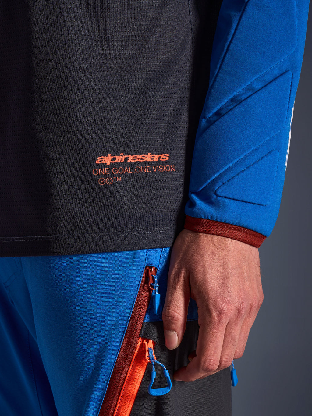 Gros plan sur le maillot Alpinestars Maxdura Dual en gris foncé et bleu royal, présentant un logo orange et le texte 'One Goal One Vision' sur le torse en maille, manche rembourrée avec panneaux géométriques renforcés, détail d'équipement technique pour la conduite d'aventure