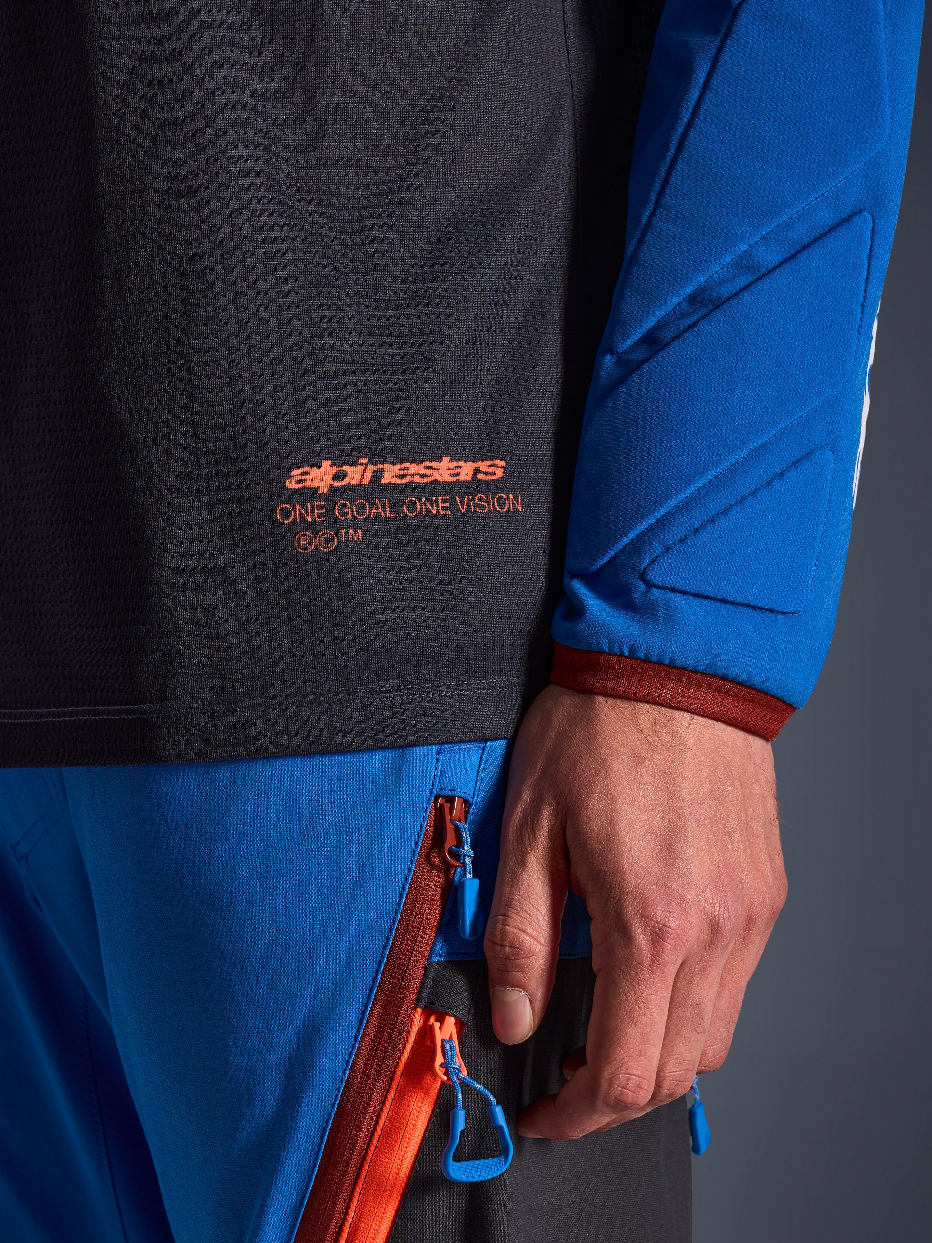 Gros plan sur le maillot Alpinestars Maxdura Dual en gris foncé et bleu royal, présentant un logo orange et le texte 'One Goal One Vision' sur le torse en maille, manche rembourrée avec panneaux géométriques renforcés, détail d'équipement technique pour la conduite d'aventure