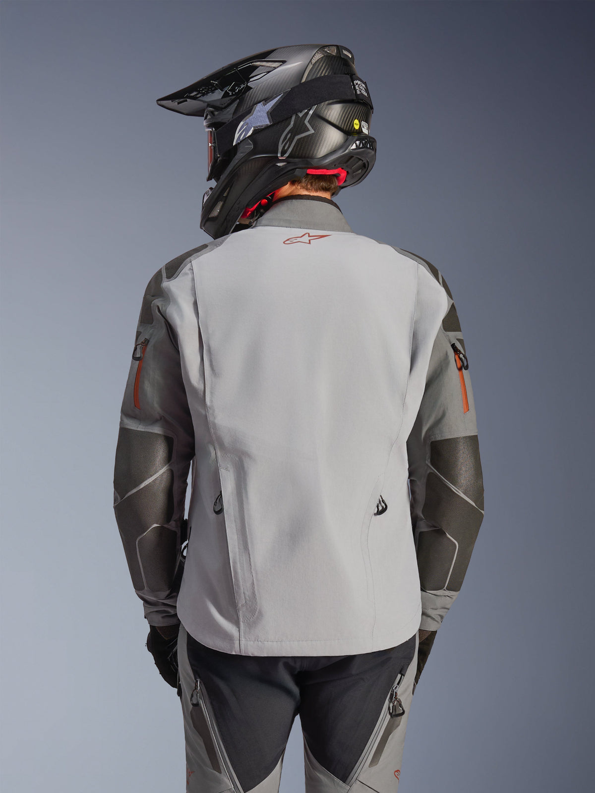 Veste Alpinestars Nazca 3L GORE-TEX PRO, veste de moto d'aventure, vue de dos, gris foncé et gris moyen, dotée de renforts Superfabric aux coudes et aux épaules, logo rouge dans le dos, portée avec un équipement d'aventure assorti et un casque en fibre de carbone.