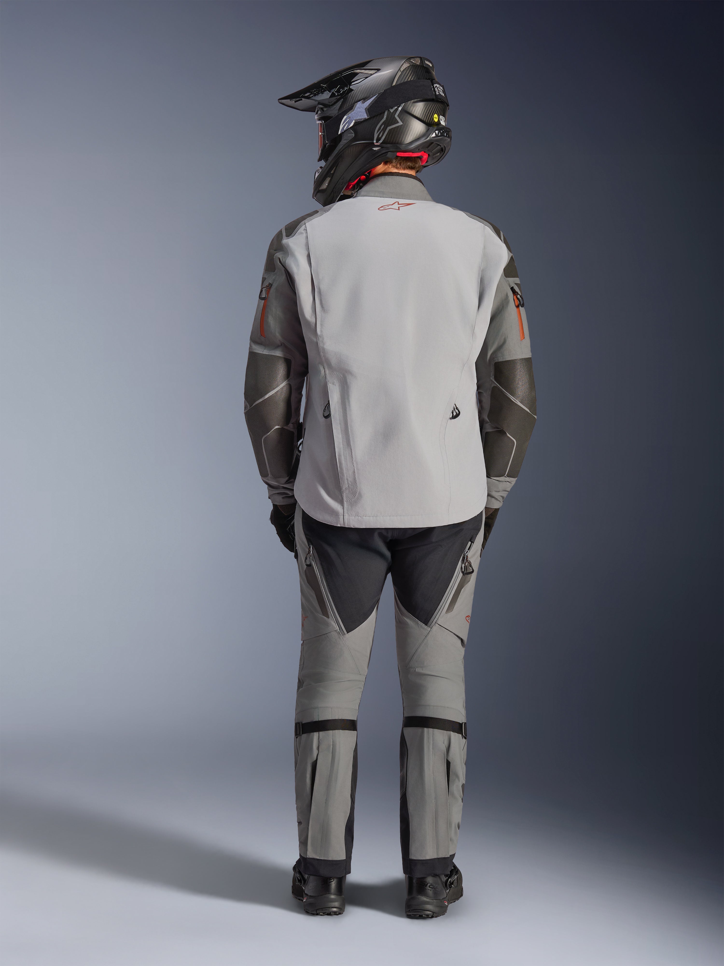 Veste Alpinestars Nazca 3L GORE-TEX PRO, veste de moto d'aventure, gris foncé et gris moyen, vue arrière d'un motard portant l'équipement complet, portée avec un pantalon assorti et un casque d'aventure noir, avec un logo rouge au dos et des renforts en Superfabric aux coudes, conception textile durable pour le touring d'aventure.