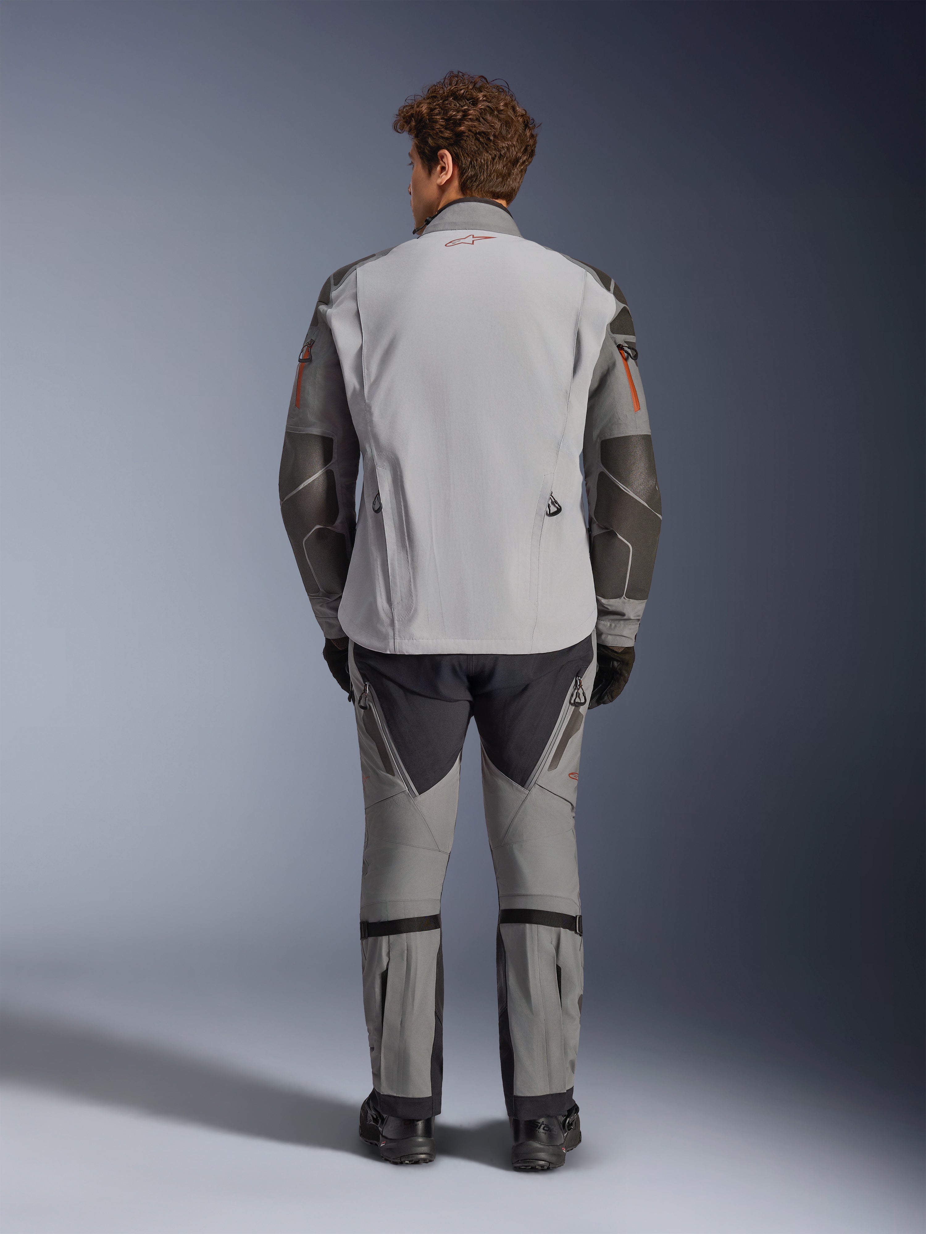 Veste Alpinestars Nazca 3L GORE-TEX PRO, vue arrière, gris foncé et gris moyen, veste de moto d'aventure avec construction GORE-TEX, logo rouge sur le col, détails orange sur les manches, portée avec un pantalon de moto assorti et des gants noirs.