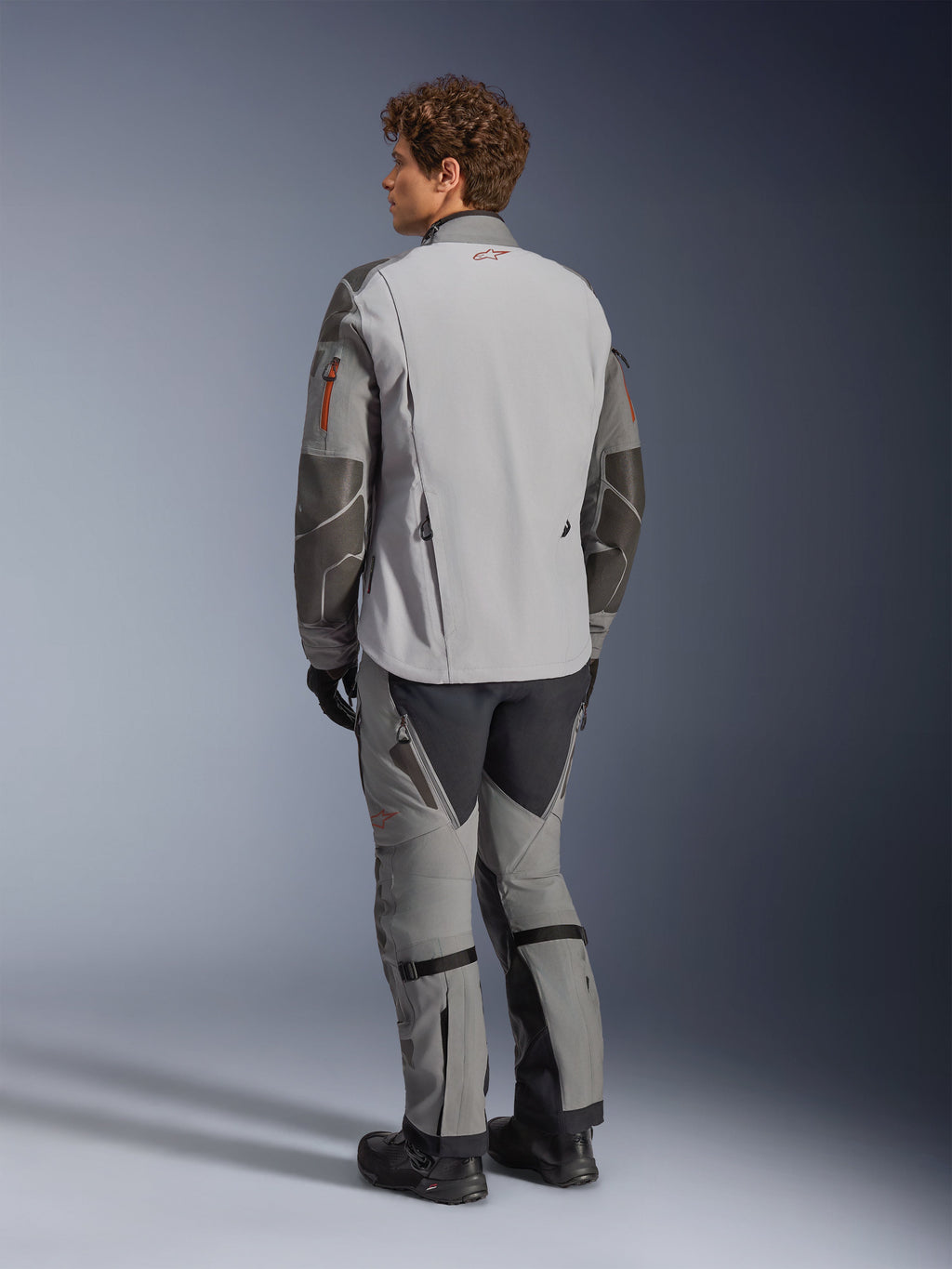 Veste Alpinestars Nazca 3L GORE-TEX PRO, veste de moto d'aventure, gris foncé et gris moyen, vue arrière portée avec un pantalon d'aventure assorti et des bottes noires, avec des renforts Superfabric gris foncé sur les manches, des touches orange sur les fermetures éclair et un petit logo rouge au col arrière.