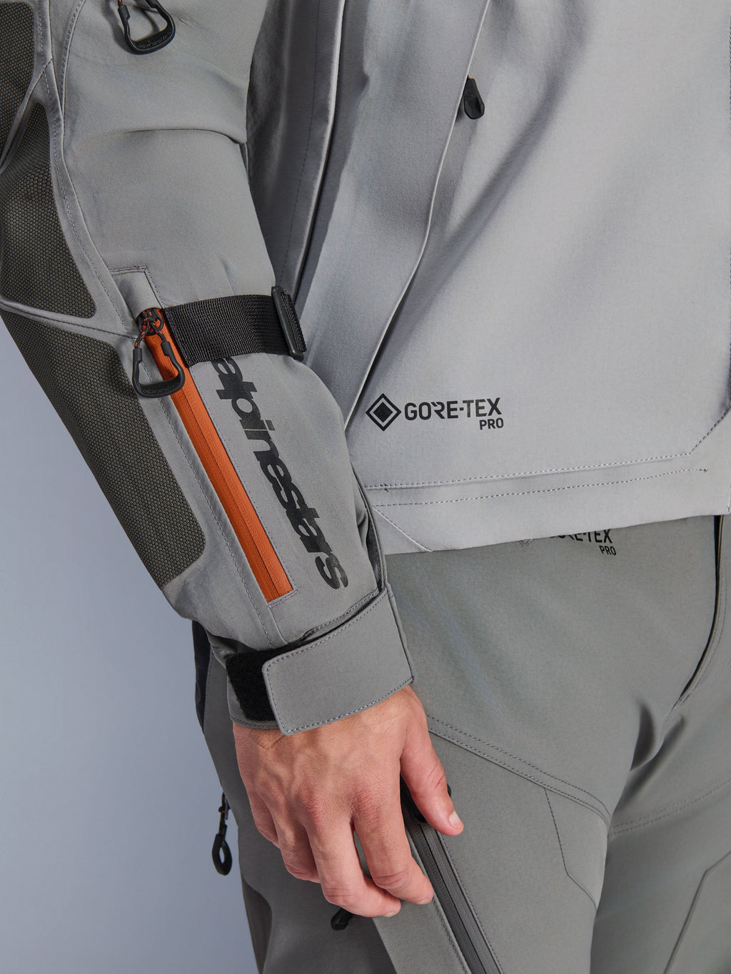 Veste Alpinestars Nazca 3L GORE-TEX PRO, veste d'aventure gris foncé et gris moyen, détail en gros plan de la manche avec marquage noir et accent de fermeture éclair orange, logo GORE-TEX PRO sur le torse, construction textile durable avec empiècement renforcé au coude