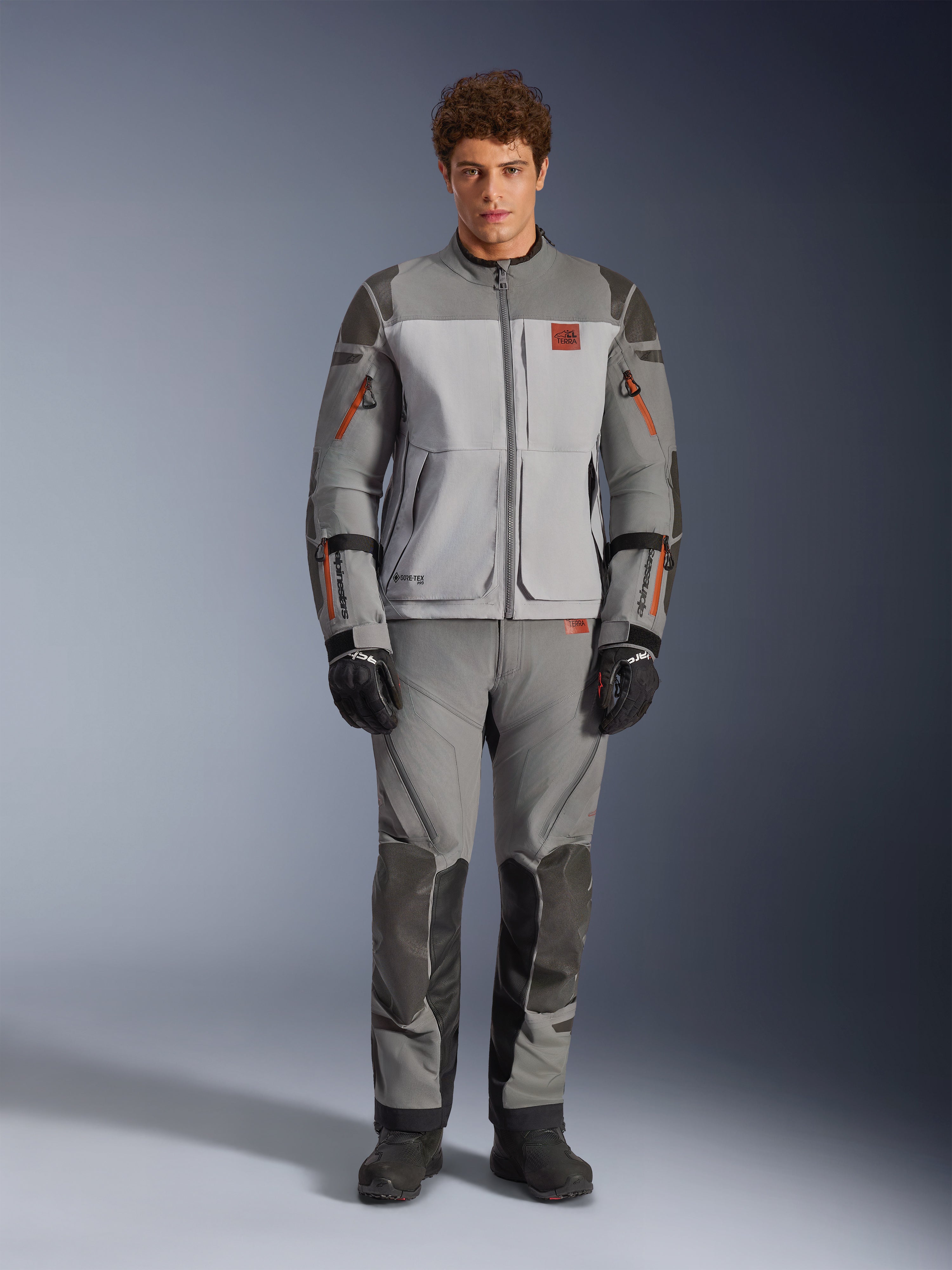 Veste Alpinestars Nazca 3L GORE-TEX PRO, veste de moto adventure, gris foncé et gris moyen, portée par un mannequin avec pantalon assorti et gants noirs, avec des accents orange sur les fermetures éclair et un logo rouge sur la poitrine, équipement de protection technique pour la conduite extrême