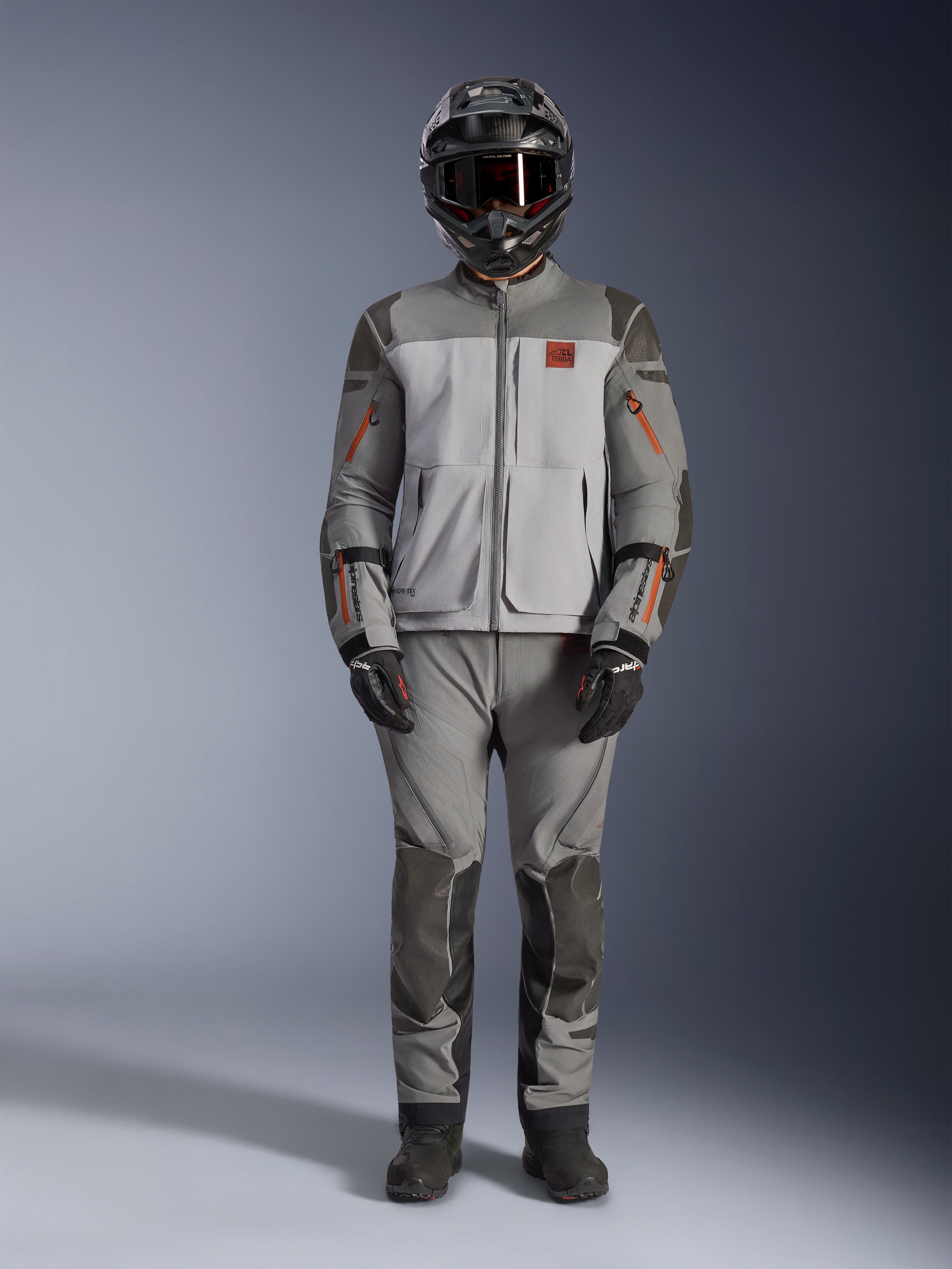 Veste Alpinestars Nazca 3L GORE-TEX PRO, veste de moto adventure, gris foncé et gris moyen avec accents orange, dotée de renforts Superfabric et de zips de ventilation à l'avant, portée avec un pantalon assorti et un casque noir, équipement de conduite technique tout-terrain