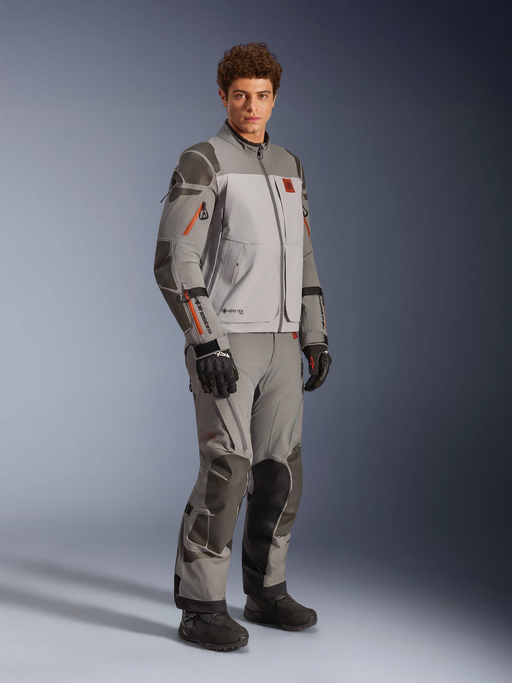 Veste Alpinestars Nazca 3L GORE-TEX PRO en Gris Foncé Gris Moyen, tenue de moto d'aventure portée avec un pantalon assorti et des gants noirs, présentant des accents de fermeture éclair orange, des renforts protecteurs en Superfabric® et le marquage GORE-TEX.