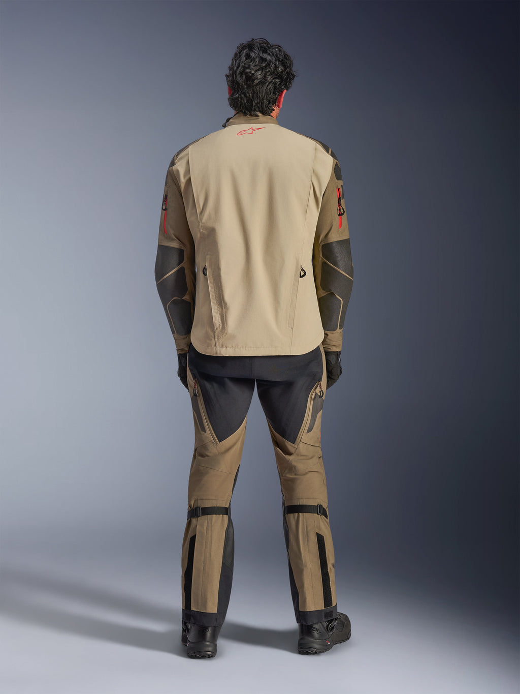 Alpinestars Nazca 3L GORE-TEX PRO Jacket in Walnut Rock Brown, back view of rider, showing Superfabric reinforcements on elbows and rear ventilation zippers, worn with matching adventure pants, black boots, and gloves, rugged technical riding apparel for extreme adventure.