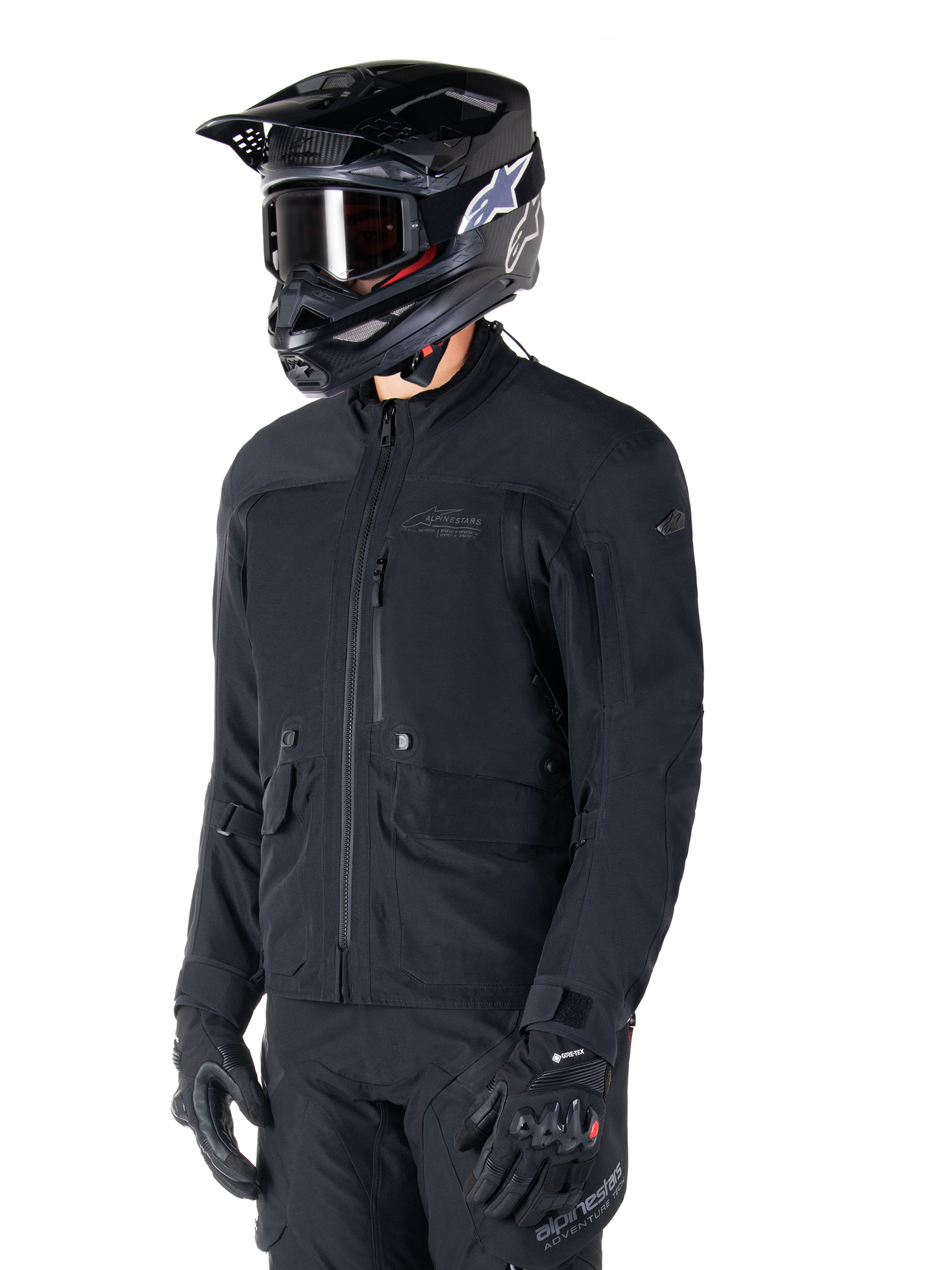 Veste Alpinestars St-7 Prime GORE-TEX, veste de moto noire, portée par un pilote avec un casque noir et des lunettes de protection, dotée de grandes poches cargo sur la poitrine et d'une coque imperméable laminée, équipement premium pour l'aventure touring et les trajets quotidiens.