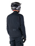 Veste Alpinestars St-7 Prime GORE-TEX, noir, vue de dos d'un pilote portant la veste avec un casque et un pantalon assortis, arborant le marquage Alpinestars et des coordonnées géographiques dans le bas du dos.