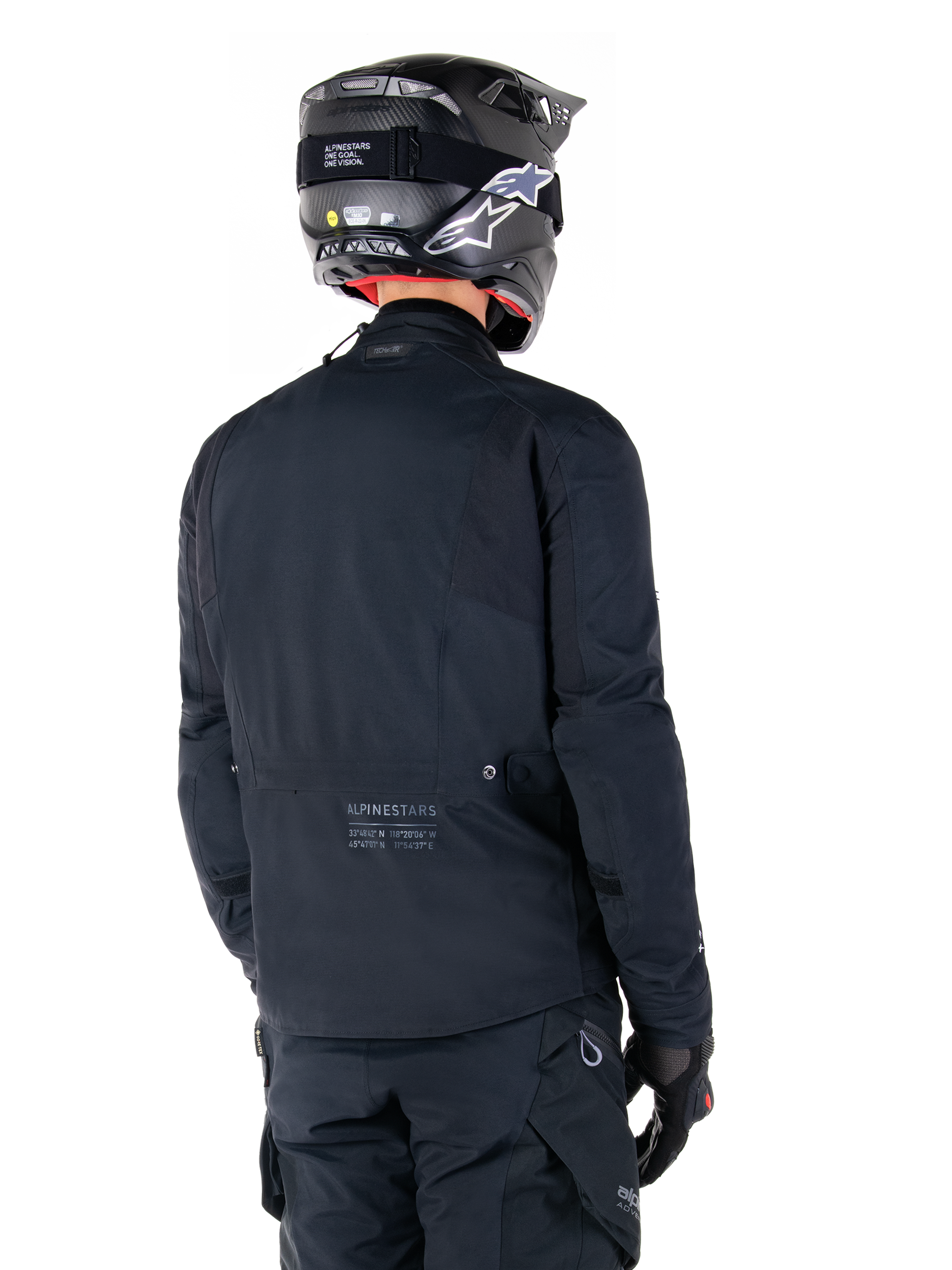 Veste Alpinestars St-7 Prime GORE-TEX, noir, vue de dos d'un pilote portant la veste avec un casque et un pantalon assortis, arborant le marquage Alpinestars et des coordonnées géographiques dans le bas du dos.