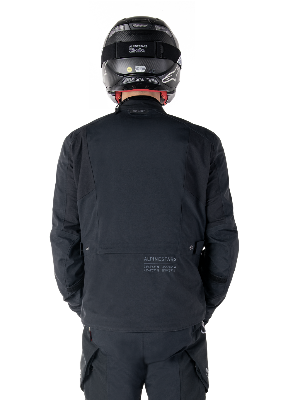 Veste Alpinestars St-7 Prime GORE-TEX, noire, vue arrière d'un pilote, avec un logo discret et des coordonnées géographiques dans le bas du dos, portée avec un casque et un pantalon noirs assortis, équipement de moto élégant et technique