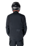 Veste Alpinestars St-7 Prime GORE-TEX, noire, vue arrière d'un pilote, avec un logo discret et des coordonnées géographiques dans le bas du dos, portée avec un casque et un pantalon noirs assortis, équipement de moto élégant et technique