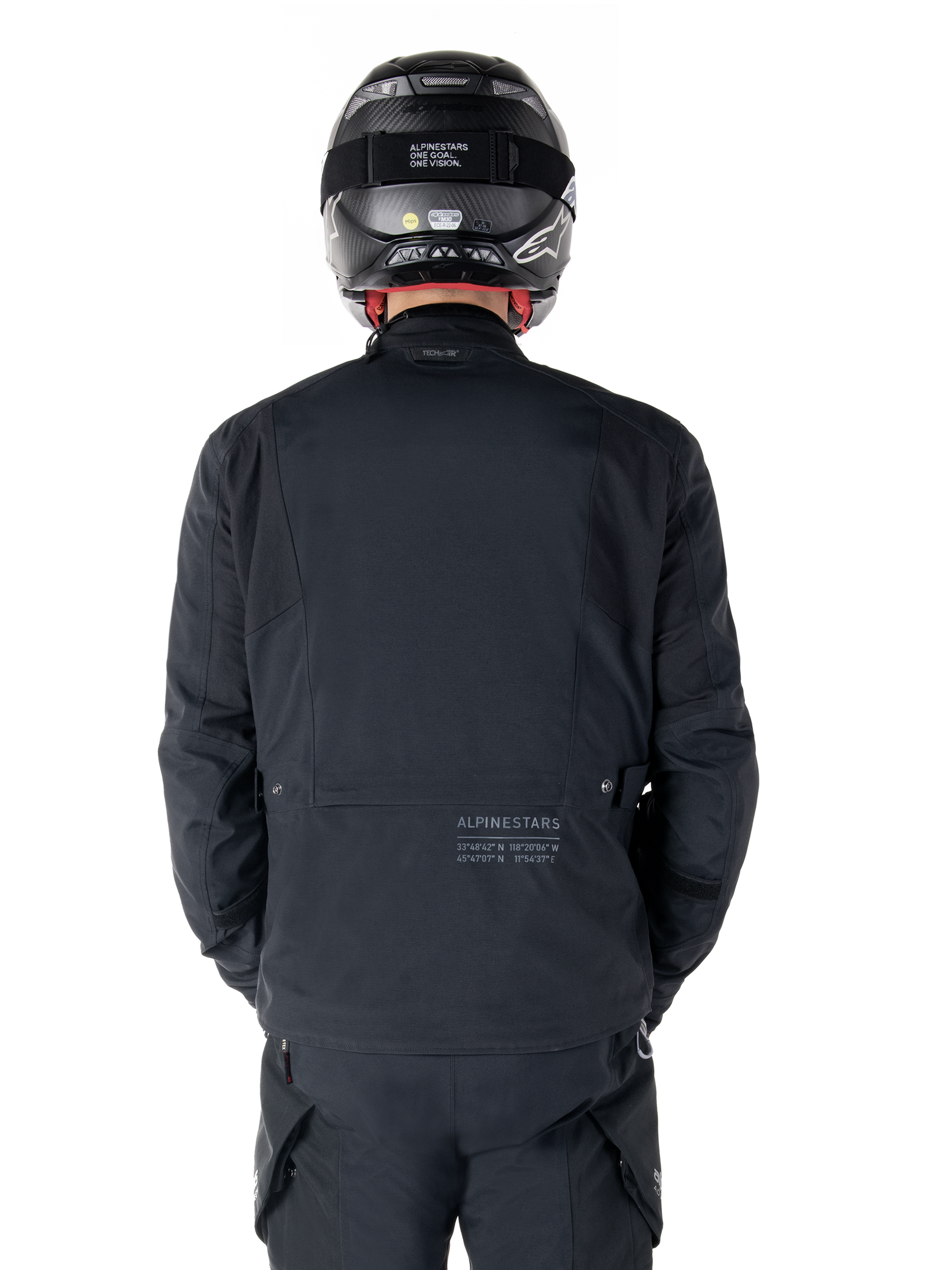Veste Alpinestars St-7 Prime GORE-TEX, noire, vue arrière d'un pilote, avec un logo discret et des coordonnées géographiques dans le bas du dos, portée avec un casque et un pantalon noirs assortis, équipement de moto élégant et technique
