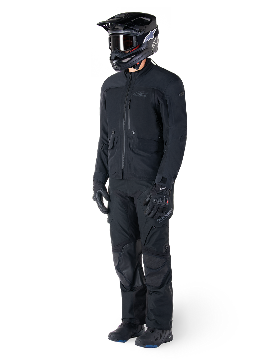 Motocycliste portant la veste Alpinestars St-7 Prime GORE-TEX noire, tenue d'aventure touring coordonnée avec pantalon noir assorti, casque noir avec lunettes et gants, présentant un design épuré et fonctionnel pour des performances par tous les temps.