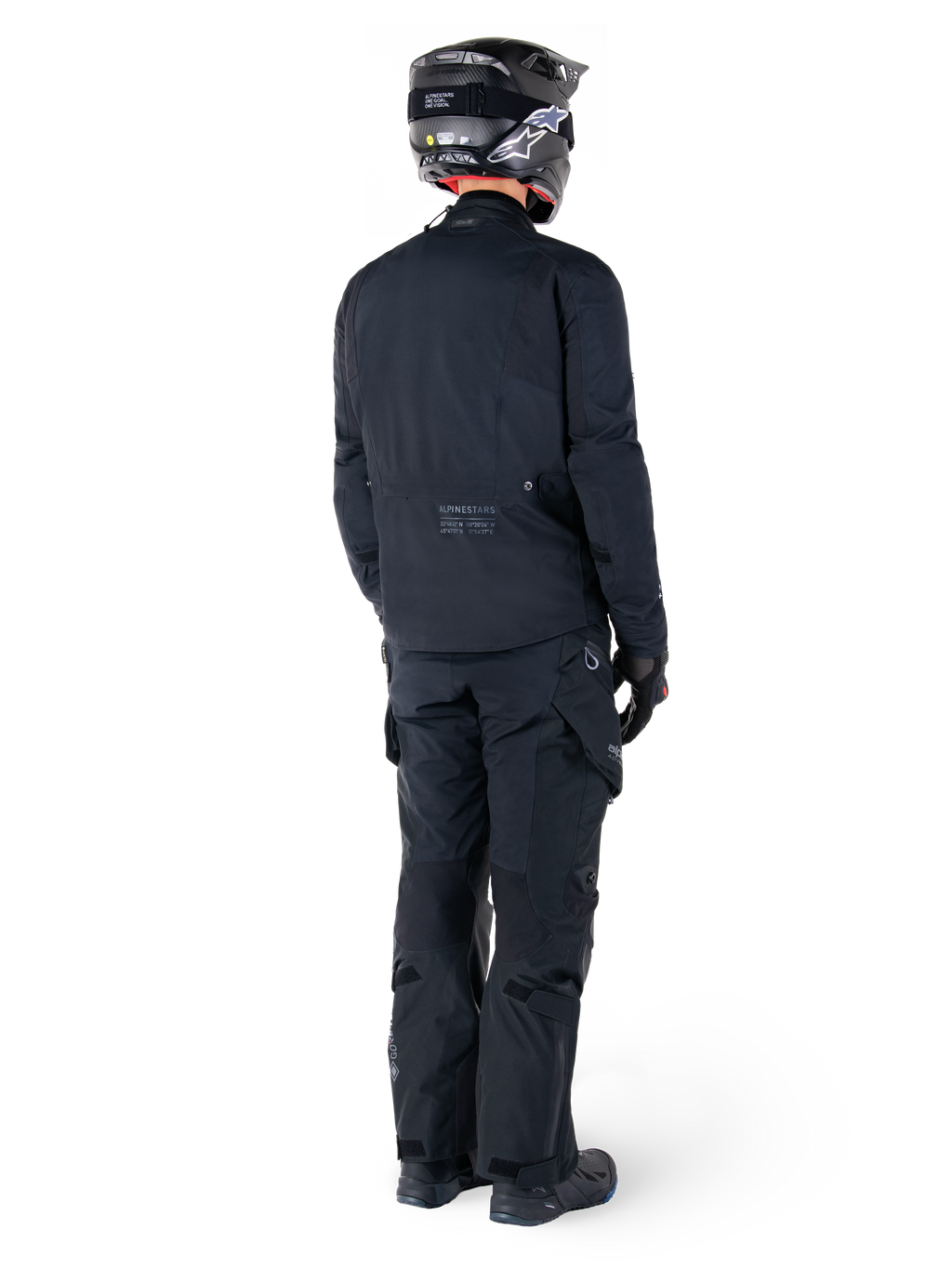 Vue arrière d'un pilote portant la veste Alpinestars St-7 Prime GORE-TEX noire, associée à un pantalon de touring noir assorti, un casque de moto noir avec logo blanc et des bottes de moto noires, équipement d'aventure protecteur complet pour le touring et les trajets quotidiens.
