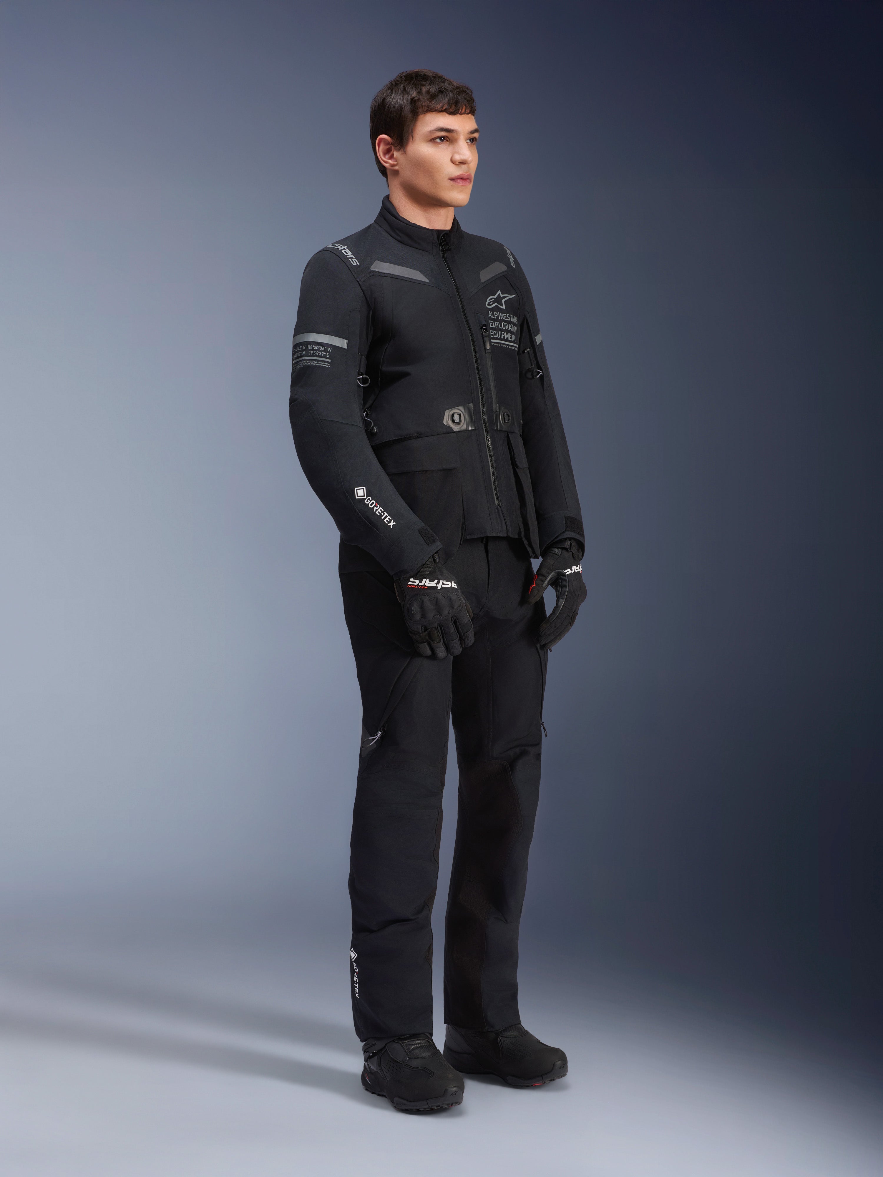 Mannequin homme portant la veste Alpinestars ST-7 2L GORE-TEX en Noir Gris Foncé, veste de moto adventure touring, pantalon et gants noirs assortis, vue de trois-quarts en pied, avec logo GORE-TEX et marquage Alpinestars Exploration Equipment