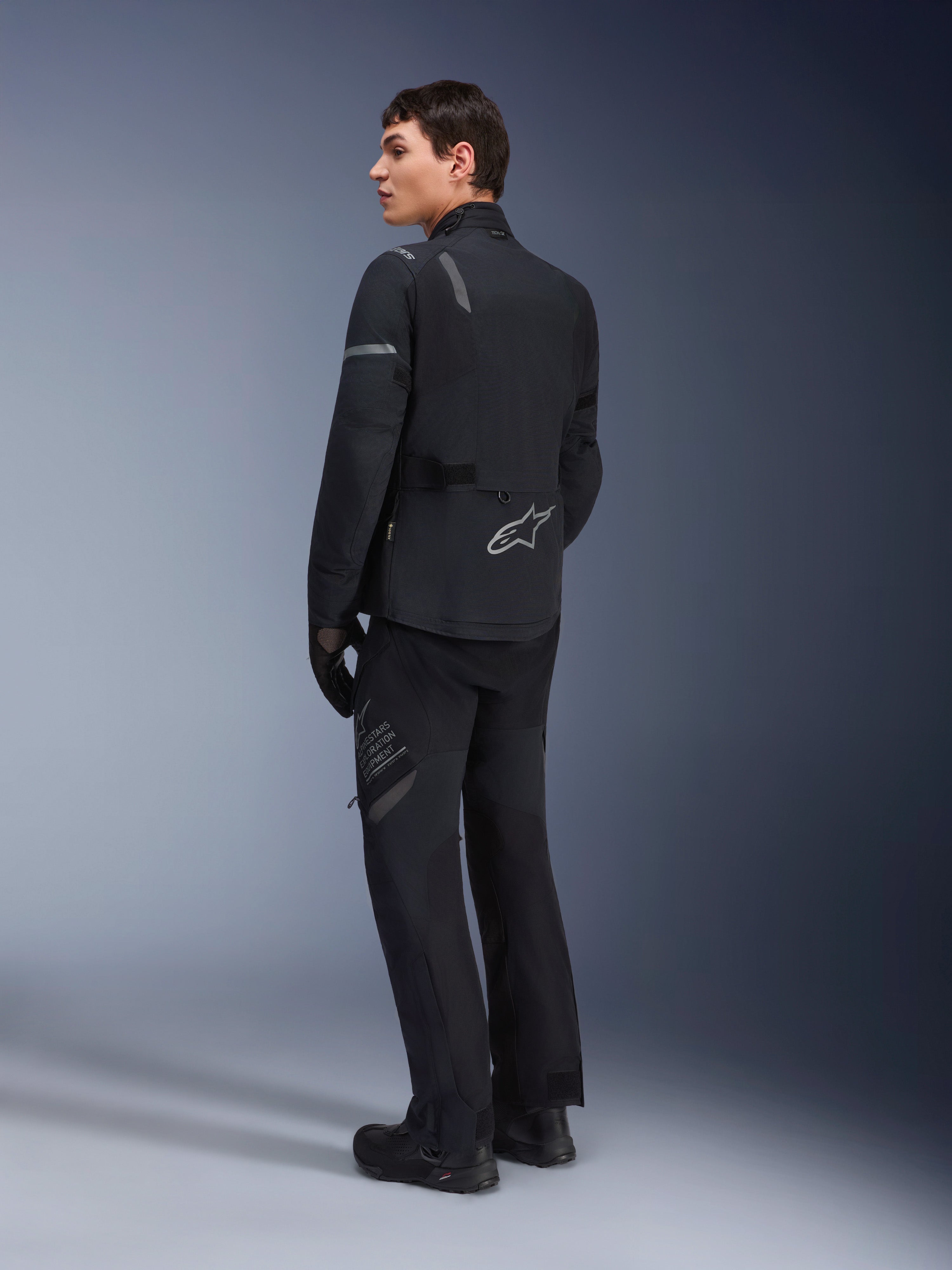 Pantalon Alpinestars ST-7 2L GORE-TEX, pantalon de touring d'aventure, noir et gris foncé, porté avec la veste ST-7 assortie et des bottes noires, arborant le marquage 'Alpinestars Exploration Equipment' sur la cuisse, conçu pour une conduite imperméable et respirante toutes saisons