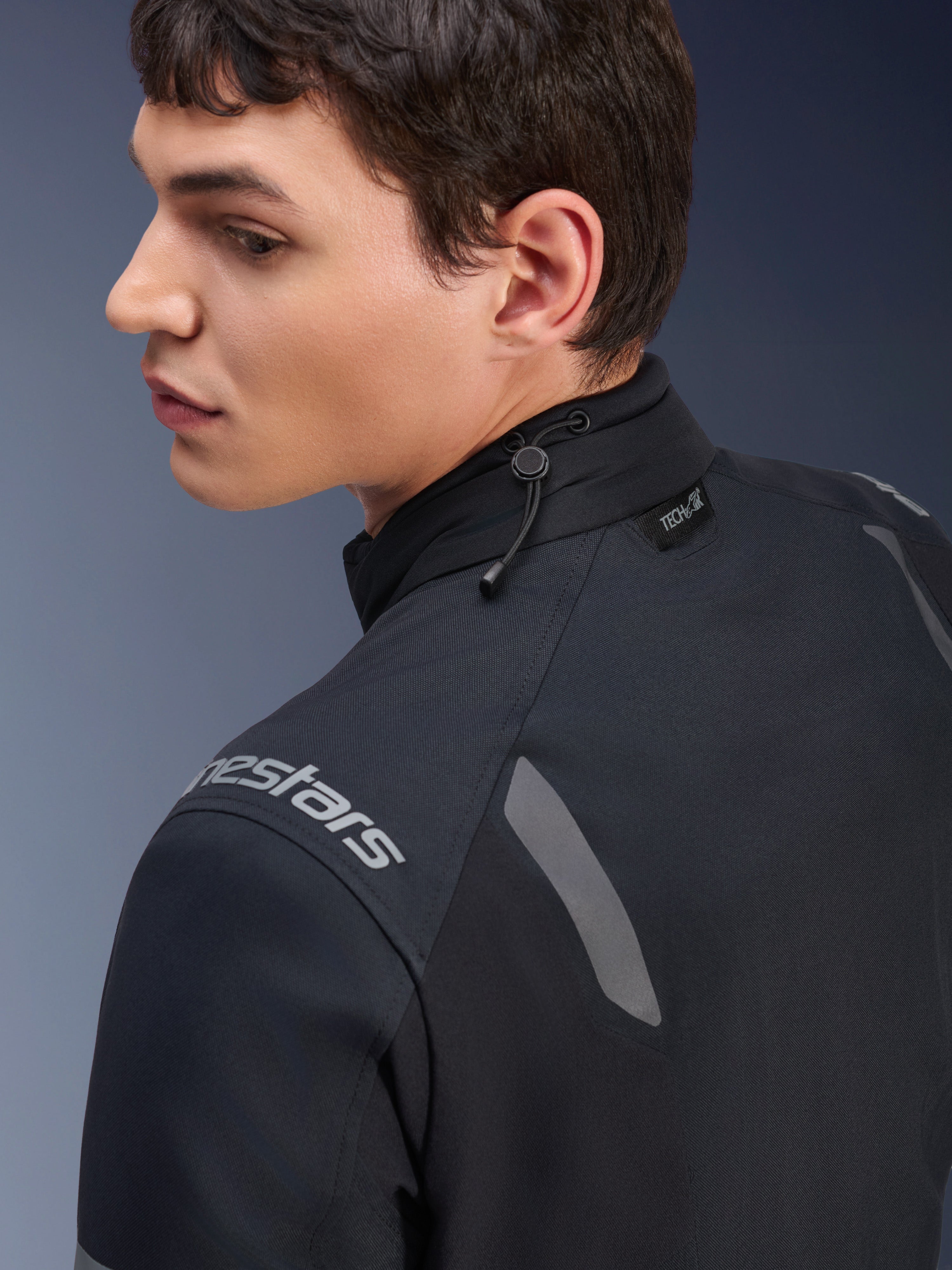 Veste Alpinestars ST-7 2L GORE-TEX, veste de moto noire et gris foncé, vue rapprochée du col montrant l'ajustement par cordon de serrage et l'étiquette Tech-Air, avec accents réfléchissants gris et logo de la marque sur l'épaule.