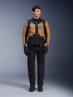 Modèle masculin portant la veste Alpinestars ST-7 2L GORE-TEX, Gobi Brown et noir, équipement de moto aventure avec pantalon et gants noirs assortis, arborant le logo Alpinestars Exploration Equipment sur la poitrine, conception imperméable fonctionnelle pour le touring sur route et hors-piste