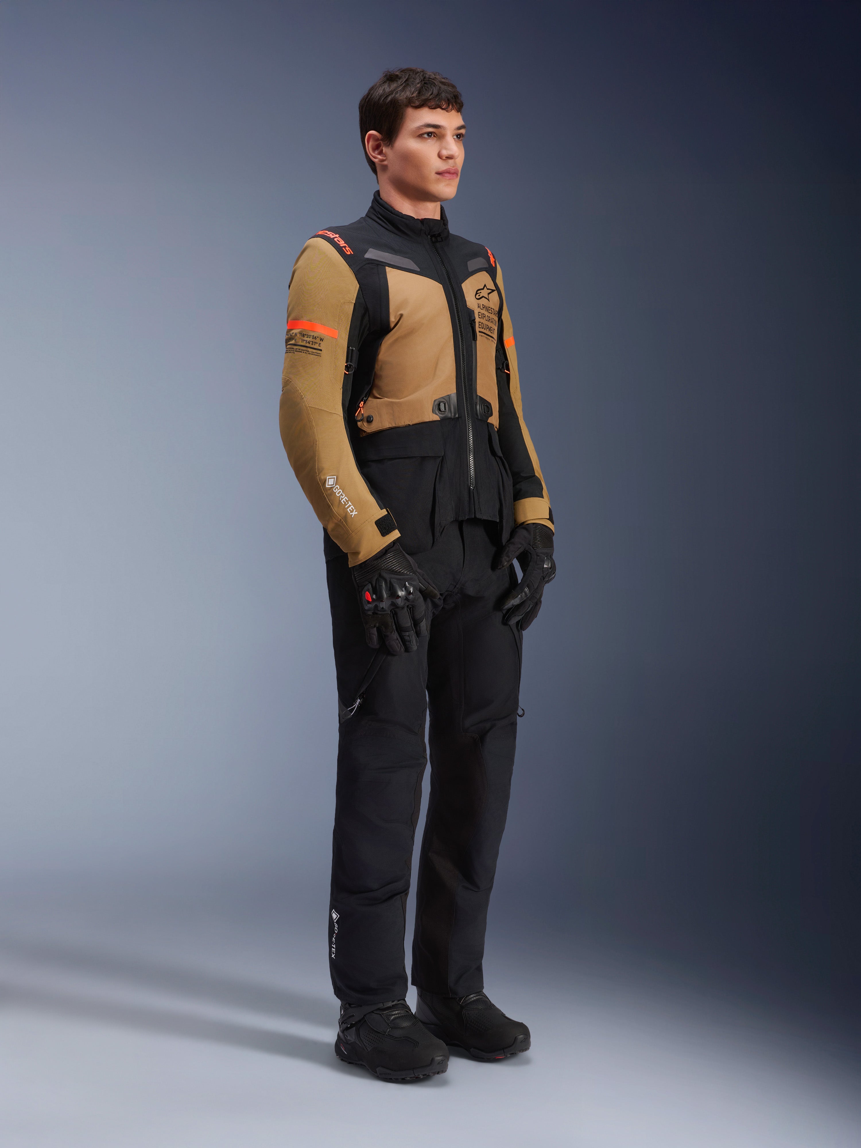 Veste Alpinestars ST-7 2L GORE-TEX, veste de moto, Brun Gobi et noir, portée par un mannequin avec un pantalon et des gants noirs assortis, avec des panneaux marron sur la poitrine et les manches, des accents techniques noirs et un logo orange, équipement de moto d'aventure-tourisme.