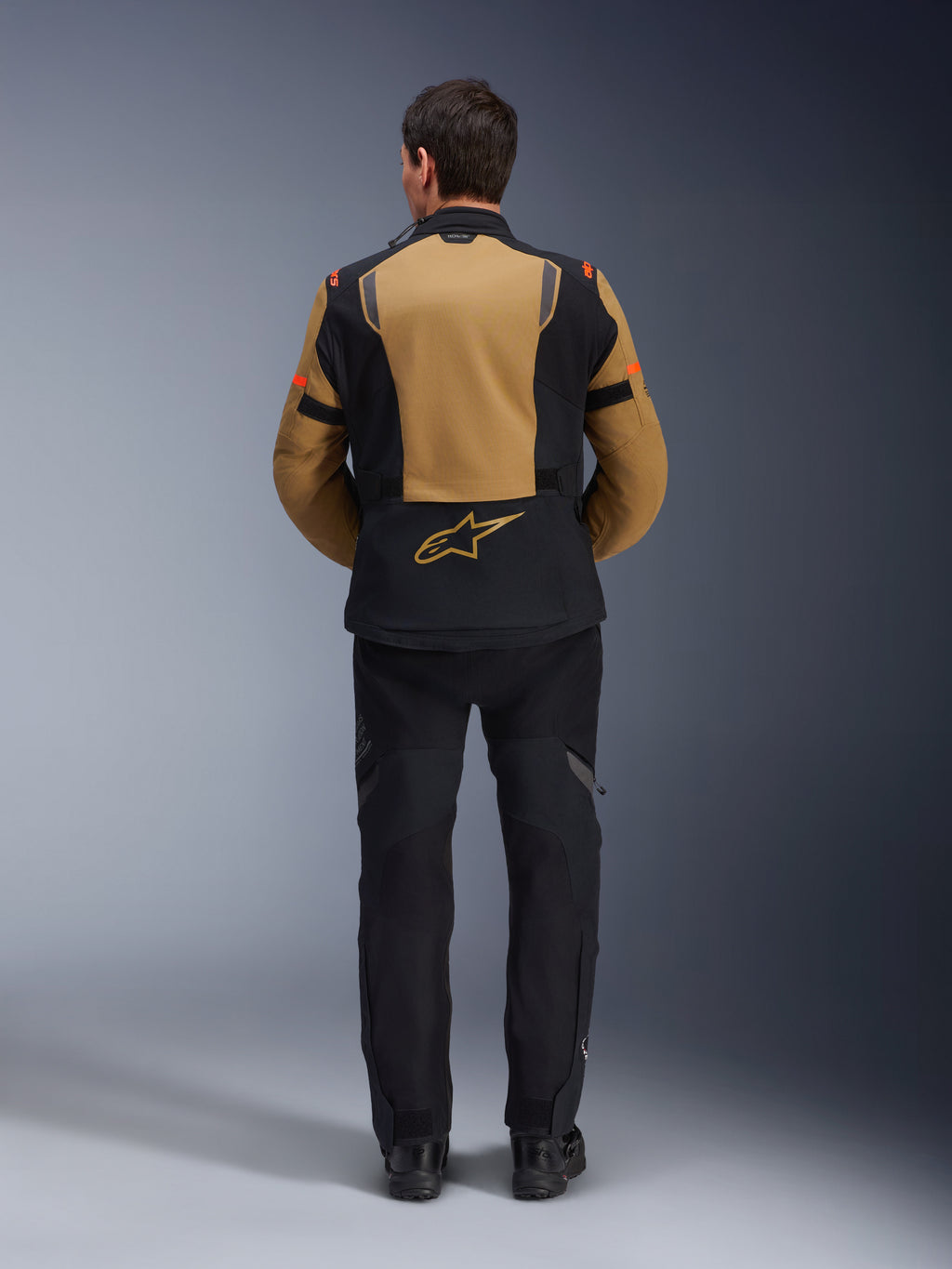 Veste Alpinestars ST-7 2L GORE-TEX, veste de moto aventure, brun Gobi et noir, vue arrière montrant les empiècements ergonomiques et le logo Alpinestars, portée avec un pantalon de moto et des bottes noirs, équipement de touring imperméable