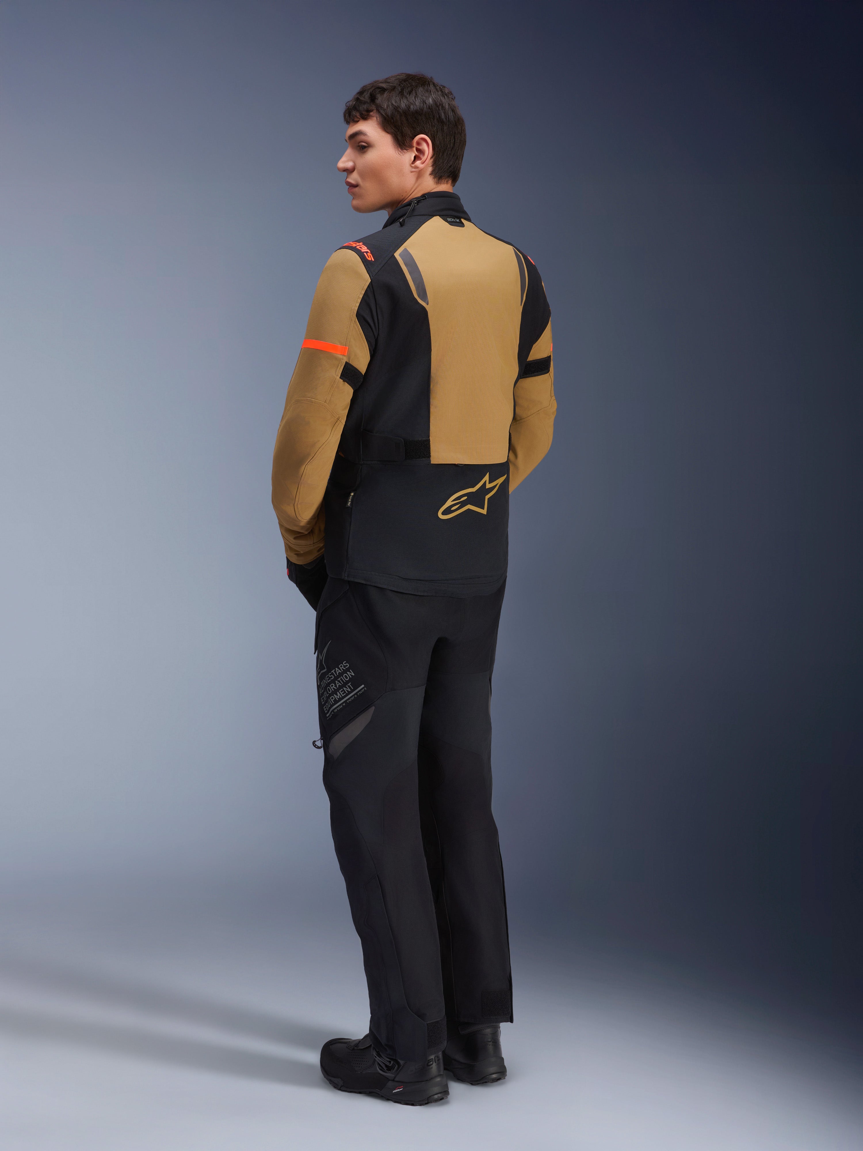 Veste Alpinestars ST-7 2L GORE-TEX, veste de moto d'aventure, brun Gobi et noir, vue arrière sur un mannequin portant un pantalon et des bottes de moto assortis, avec des accents orange sur les manches, coque laminée imperméable et logo Alpinestars, conçue pour le touring et la route