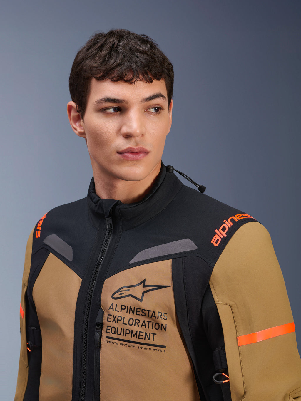 Veste Alpinestars ST-7 2L GORE-TEX, brun Gobi et noir, portée par un mannequin homme dans un studio, avec marquage « ALPINESTARS EXPLORATION EQUIPMENT » sur la poitrine, logos orange sur les épaules et textile technique imperméable pour le pilotage d'aventure.