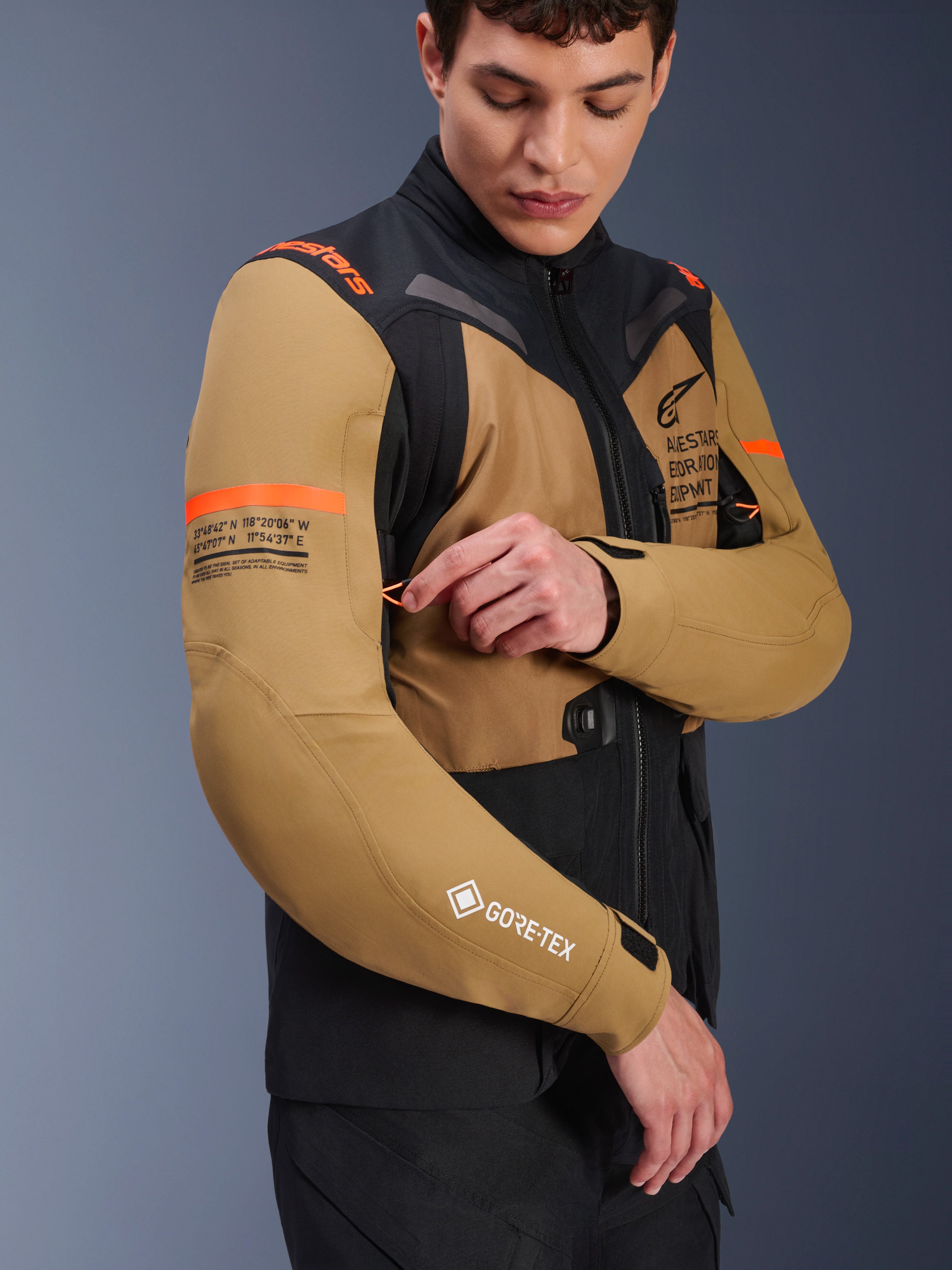 Veste Alpinestars ST-7 2L GORE-TEX en marron Gobi et noir, veste de moto adventure-touring portée par un mannequin ajustant une aération sur la manche, avec des accents orange, des graphismes de coordonnées et le logo GORE-TEX