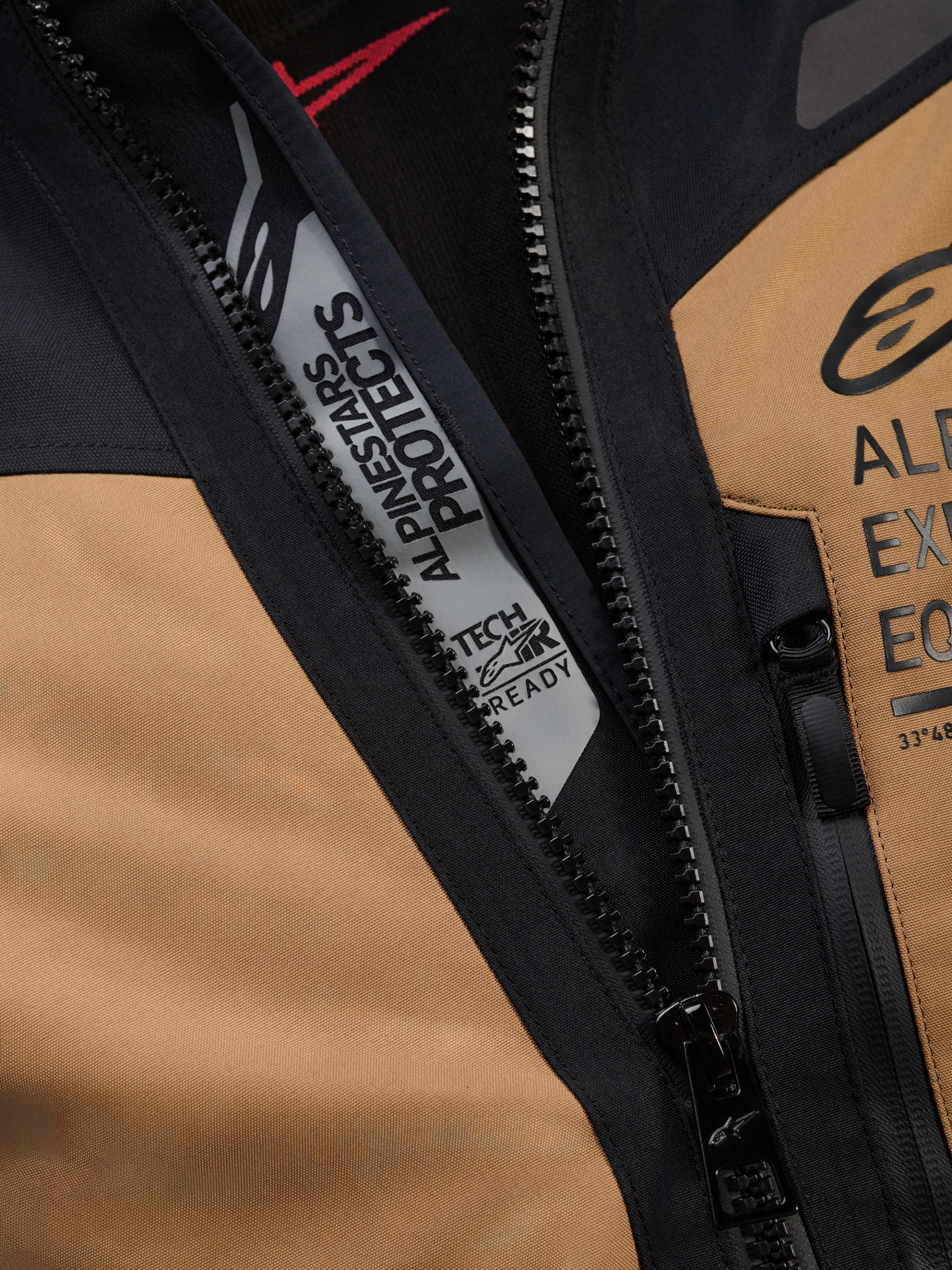 Gros plan détaillé de la veste Alpinestars ST-7 2L GORE-TEX en Marron Gobi et noir, arborant les marquages « ALPINESTARS PROTECTS » et « TECH-AIR READY » à l'intérieur de la fermeture éclair principale, mettant en évidence la construction textile robuste et les logos de poitrine.