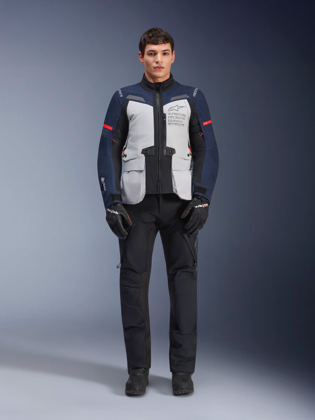 Alpinestars ST-7 2L GORE-TEX Jacket, Adventure Touring Motorcycle Jacket, Ice Gray Dark Blue and Black, worn with black pants and gloves, featuring large front cargo pockets, red accents on the sleeves, and a laminated GORE-TEX shell for waterproof performance.