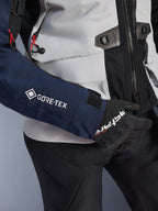 Alpinestars ST-7 2L GORE-TEX Jacket, close-up of dark blue sleeve with white GORE-TEX logo, ice gray torso with black detailing and red cord accents, worn with black Alpinestars gloves and pants, adventure touring motorcycle gear
