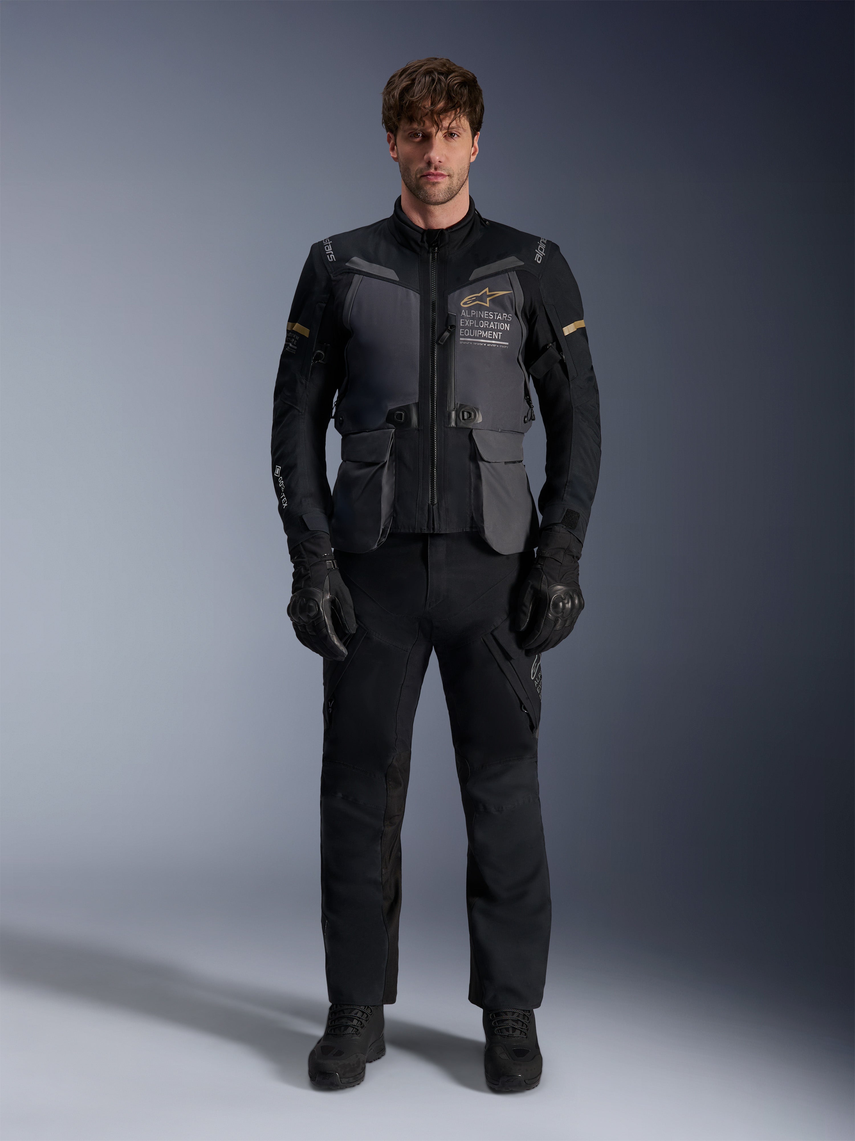 Male model wearing the Alpinestars ST-7 2L GORE-TEX Jacket in Deep Gray Black Dark Khaki, featuring 'ALPINESTARS EXPLORATION EQUIPMENT' branding on the chest, paired with matching black riding pants, gloves, and boots, studio front view.