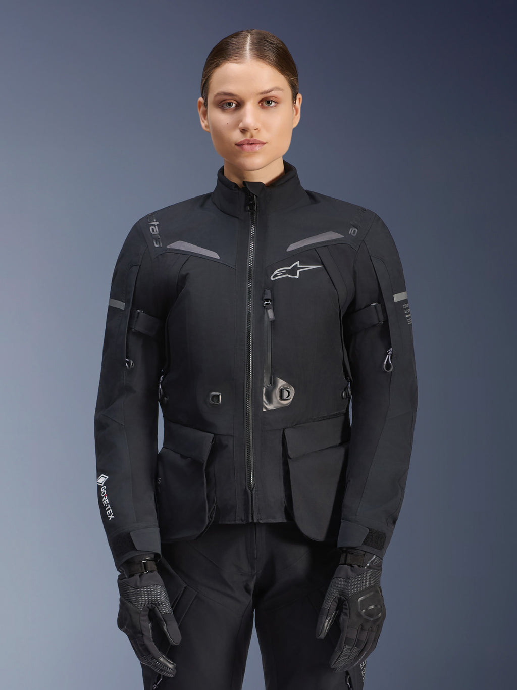 Femme portant une veste Alpinestars Stella ST-7 2L GORE-TEX, noire avec accents gris foncé, veste de moto touring avec grandes poches cargo et col haut, associée à des gants et un pantalon noirs, équipement d'aventure imperméable