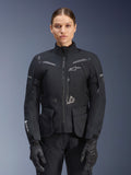 Femme portant une veste Alpinestars Stella ST-7 2L GORE-TEX, noire avec accents gris foncé, veste de moto touring avec grandes poches cargo et col haut, associée à des gants et un pantalon noirs, équipement d'aventure imperméable