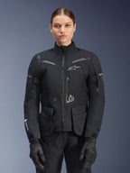 Femme portant une veste Alpinestars Stella ST-7 2L GORE-TEX, noire avec accents gris foncé, veste de moto touring avec grandes poches cargo et col haut, associée à des gants et un pantalon noirs, équipement d'aventure imperméable