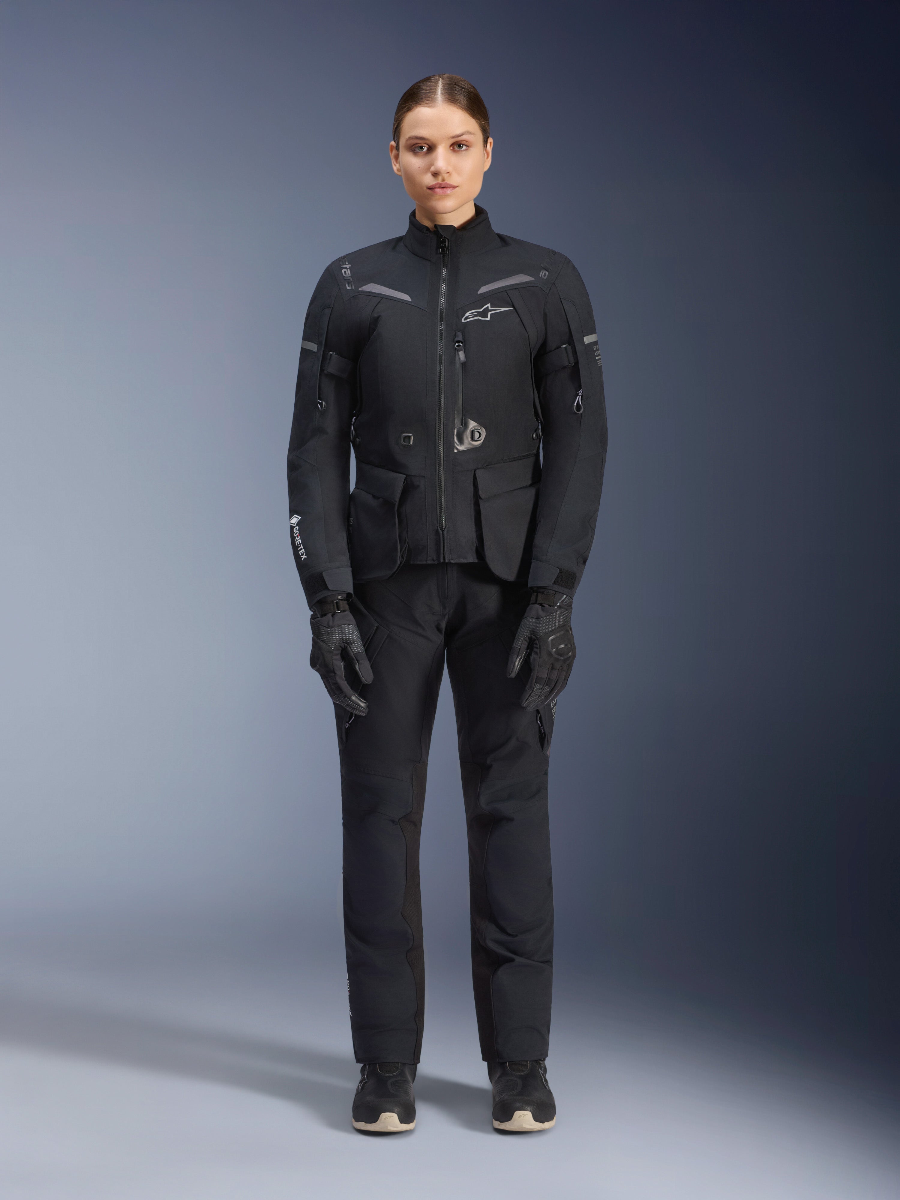 Motarde portant la veste et le pantalon Alpinestars Stella ST-7 2L GORE-TEX, tenue de touring moto noire pour femme, équipement technique imperméable et respirant, avec gants et bottes assortis, protection complète avec éléments réfléchissants pour le pilotage d'aventure