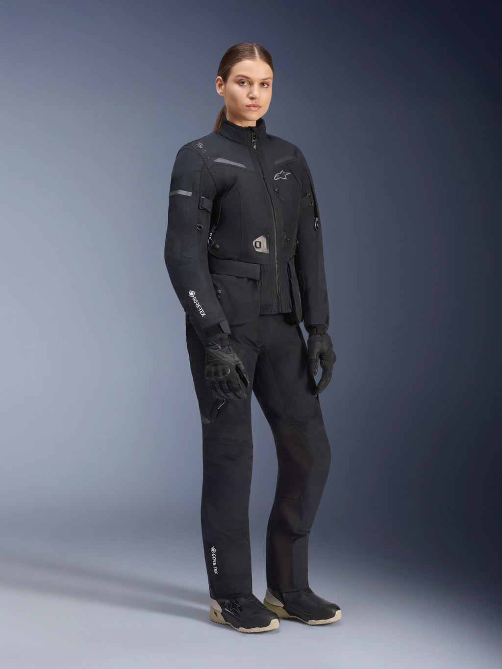 Veste Alpinestars Stella ST-7 2L GORE-TEX pour femme, noire, portée avec un pantalon de moto et des gants assortis, équipement d'aventure et de road-touring avec marquage GORE-TEX laminé sur la manche et coupe féminine ajustée.
