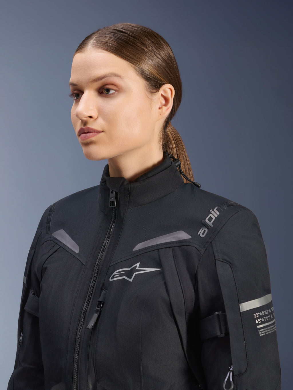 Veste Stella ST-7 2L GORE-TEX Alpinestars Femme, noire, avec logo sur la poitrine et coordonnées techniques sur le bras, design textile imperméable à col montant pour le touring à moto, portrait en studio d'une motarde.