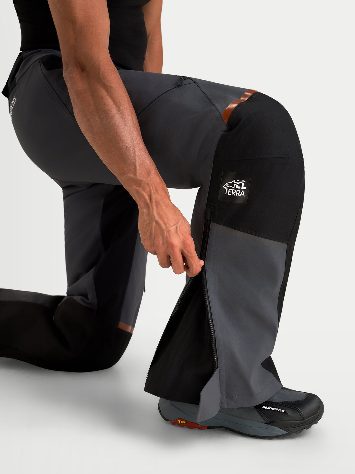 Alpinestars St-7 V2 2L Gore-Tex Pants, men's adventure touring motorcycle pants in deep gray and black, close-up view of a model kneeling and demonstrating the lower-leg zipper opening and All Terra branding.