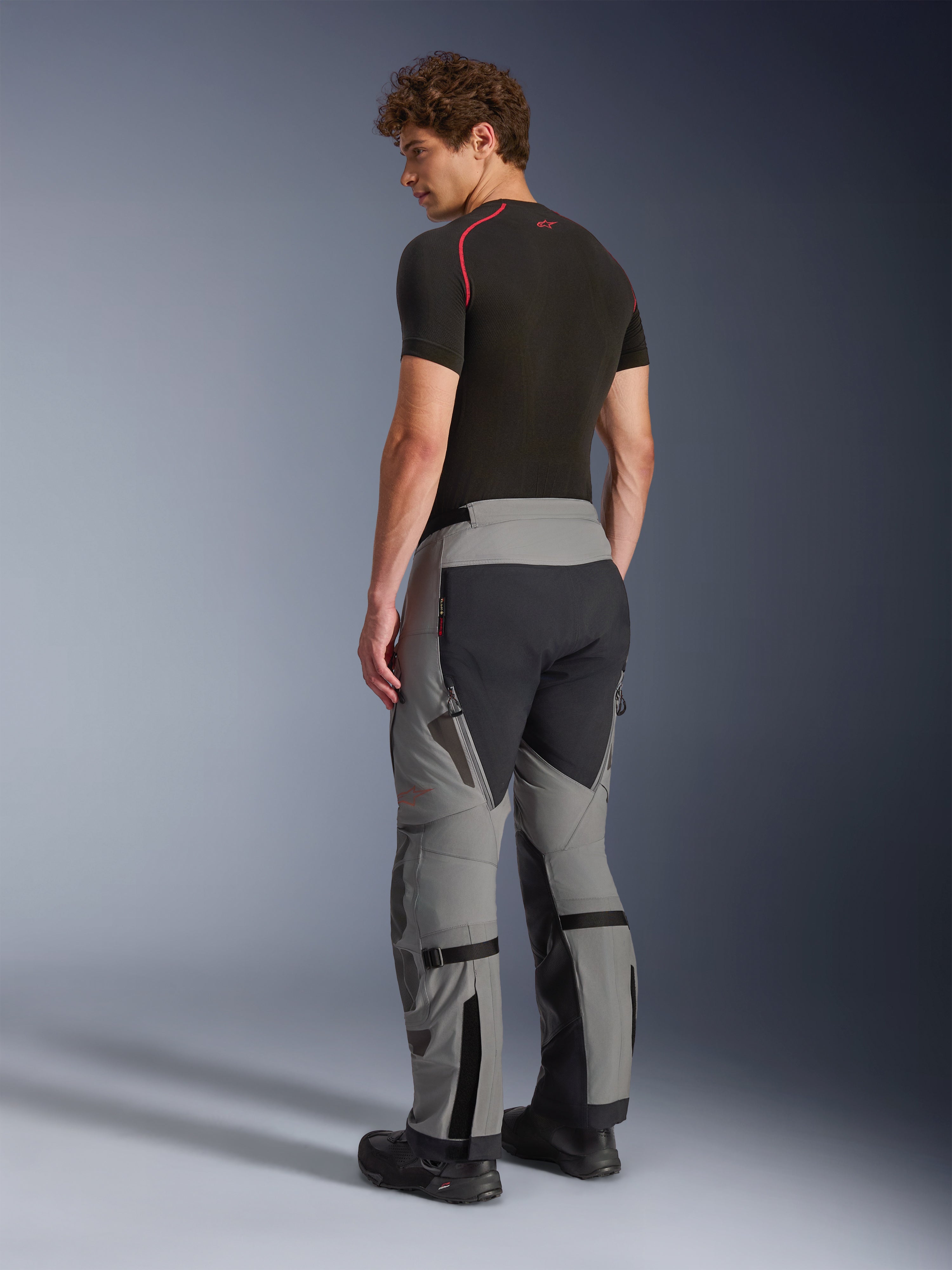 Pantalon Alpinestars Nazca 3L GORE-TEX PRO, pantalon de moto aventure, gris foncé et noir, vue arrière du mannequin portant une couche de base noire et des bottes, montrant les panneaux techniques, les zips de ventilation et les ajusteurs de volume aux mollets.