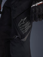 Gros plan sur le pantalon Alpinestars ST-7 2L GORE-TEX noir, vue détaillée de la grande poche cargo de cuisse avec marquage gris Alpinestars Exploration Equipment, logo étoile et coordonnées géographiques, construction textile haute performance pour le touring d'aventure.