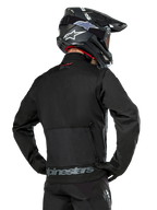 Vue arrière d'un pilote portant la veste Alpinestars Pro-Dura en noir, veste d'enduro durable en textile ripstop, dotée d'une grande poche cargo arrière avec marquage Alpinestars gris sur le bas du dos, associée à un casque de motocross noir.