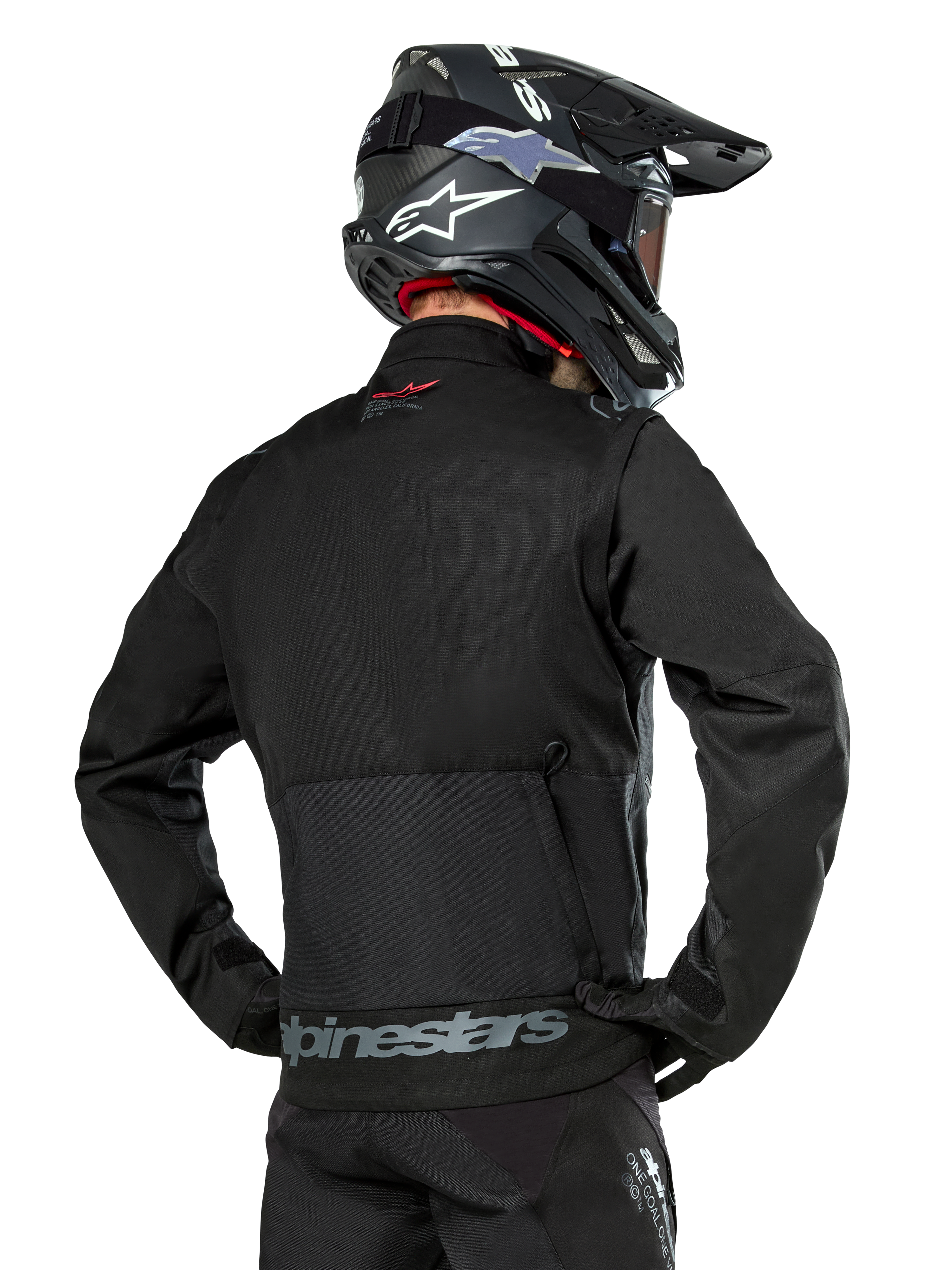 Vue arrière d'un pilote portant la veste Alpinestars Pro-Dura en noir, veste d'enduro durable en textile ripstop, dotée d'une grande poche cargo arrière avec marquage Alpinestars gris sur le bas du dos, associée à un casque de motocross noir.