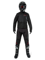 Veste Alpinestars Pro-Dura, veste d'enduro tout-terrain noire, présentée sur un modèle avec un équipement complet comprenant un casque noir avec des lunettes rouges, un pantalon noir avec des accents rouges, des bottes noires et blanches et des gants noirs, design robuste inspiré du rallye pour la conduite sur sentier