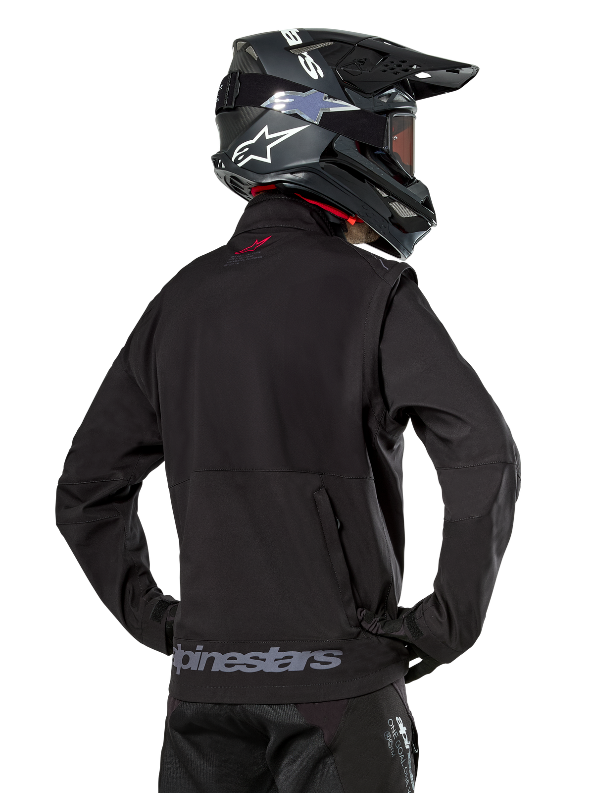 Veste Softshell Alpinestars Lite-Dura, noire, vue de dos portée par un pilote, avec marquage Alpinestars gris sur l'ourlet arrière et un petit logo rouge sur le col, associée à un casque de motocross noir, conçue pour l'enduro et la conduite tout-terrain.
