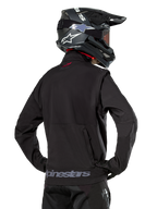 Veste Softshell Alpinestars Lite-Dura, noire, vue de dos portée par un pilote, avec marquage Alpinestars gris sur l'ourlet arrière et un petit logo rouge sur le col, associée à un casque de motocross noir, conçue pour l'enduro et la conduite tout-terrain.