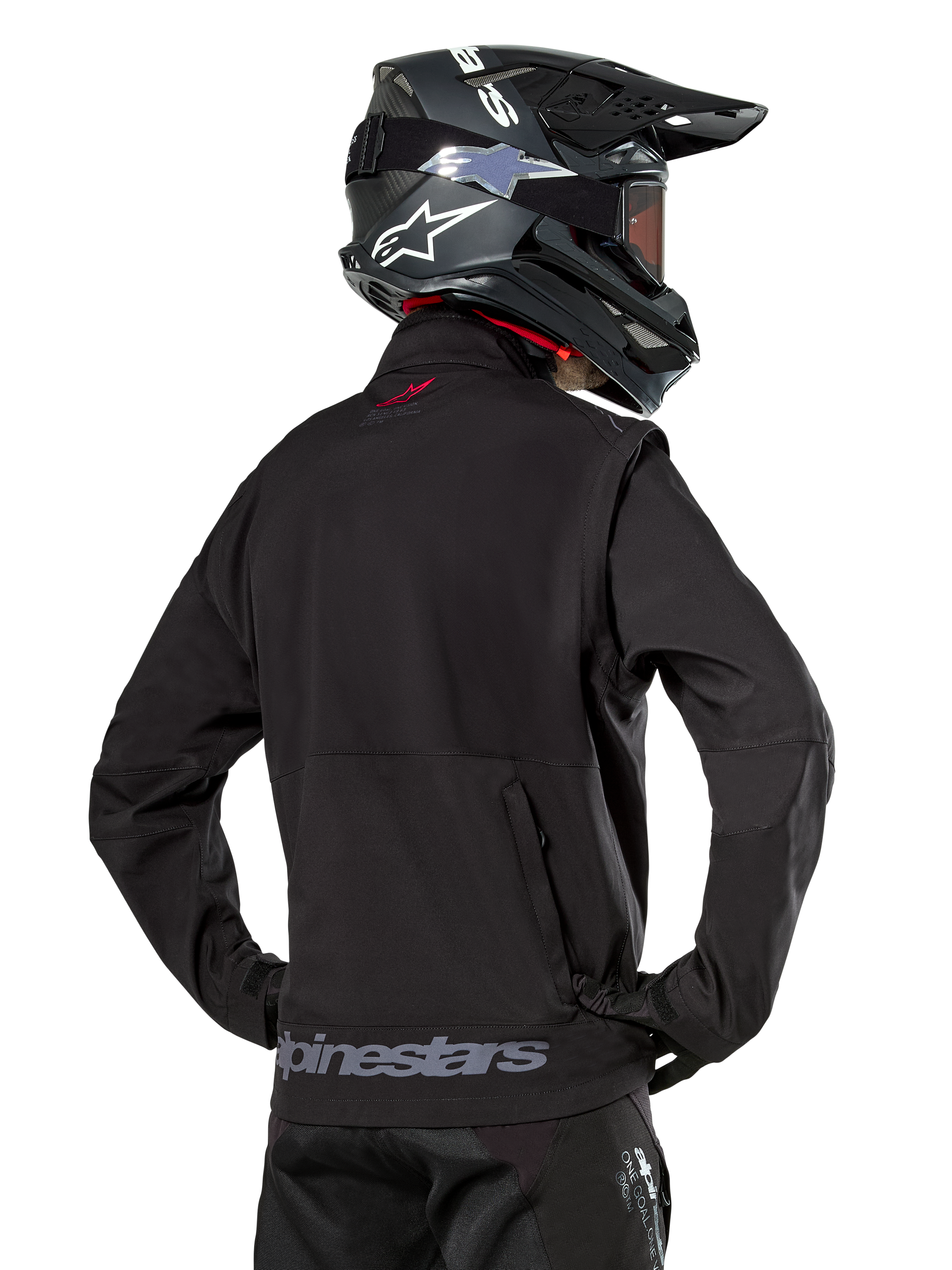 Veste Softshell Alpinestars Lite-Dura, noire, vue de dos portée par un pilote, avec marquage Alpinestars gris sur l'ourlet arrière et un petit logo rouge sur le col, associée à un casque de motocross noir, conçue pour l'enduro et la conduite tout-terrain.